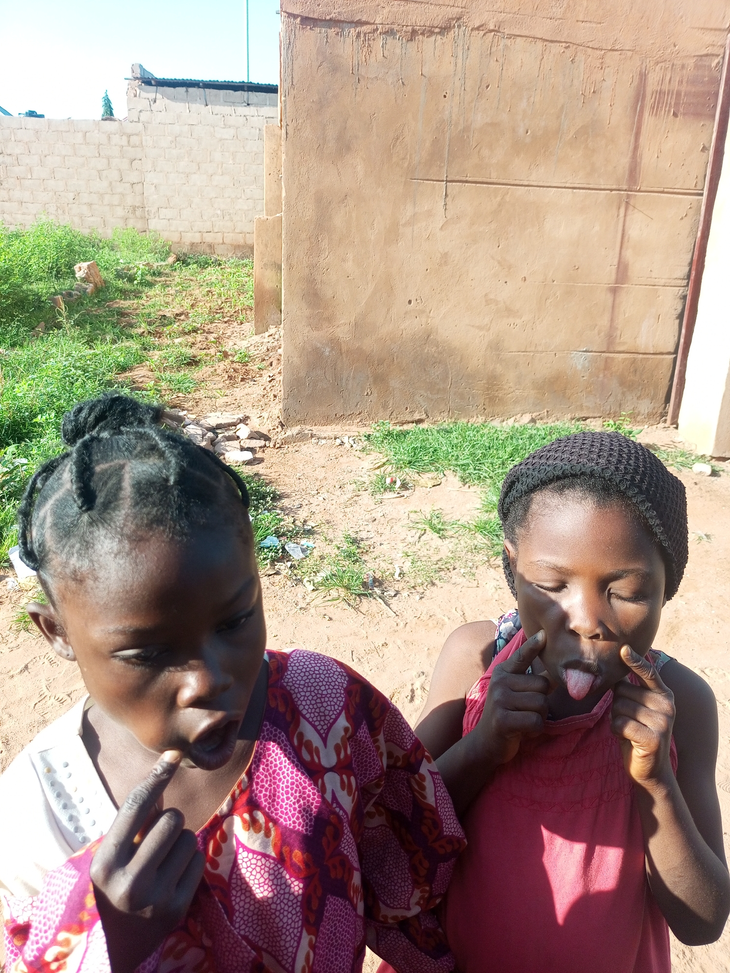 In the city of Tempure, Nigeria, two young girls are captured in a moment of playful expression against a backdrop of rudimentary urban architecture. The girl on the left, with intricately braided hair, wears a patterned garment of pink and maroon. Her mouth is open as she places a finger to her lips, her eyes focused downwards. To her right, another girl dons a red dress and a dark knitted cap. Her eyes are closed as she pulls at the corners of her mouth with her fingers, sticking out her tongue. The ground beneath them is composed of dry earth and scattered stones, with sparse green vegetation growing around. Behind them, a rough, sand-colored wall dominates the scene, with a brick wall visible in the distance to the left. The sky above is a clear, pale blue, indicating a bright, sunny day. The overall impression is one of casual, everyday life in a community characterized by its unpretentious surroundings.
