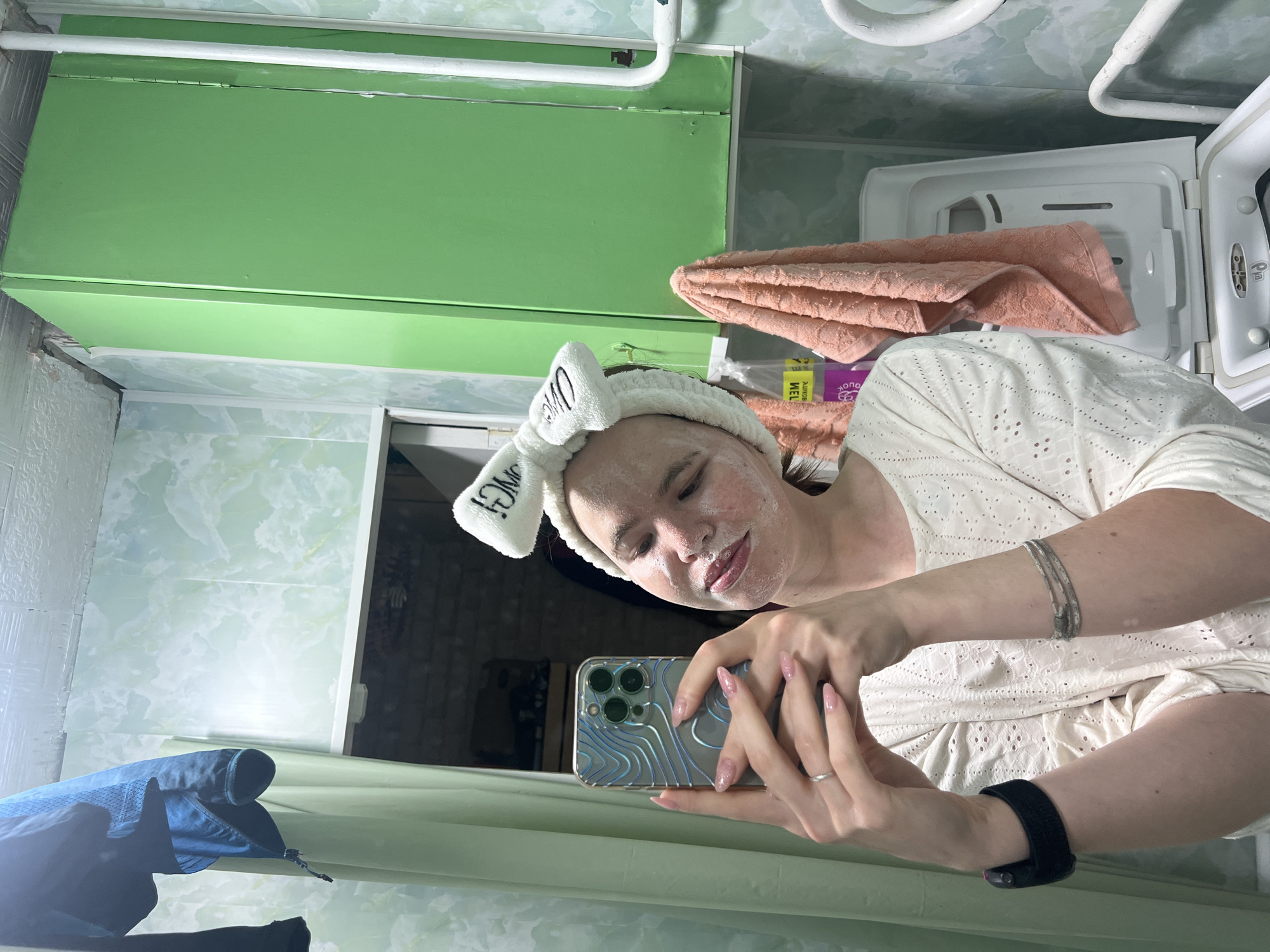 A woman is taking a selfie in what appears to be a bathroom. She's wearing a white headband with a bow and lettering, and a white, lace-like top. Her face has a cream or mask applied. She holds an iPhone with a patterned case. A black fitness tracker is visible on her wrist.
The bathroom has a green cabinet, a white washing machine, and an assortment of towels. The walls are decorated with a light green marble-like pattern. The bathroom appears to be in Chaikovsky, Russia.
