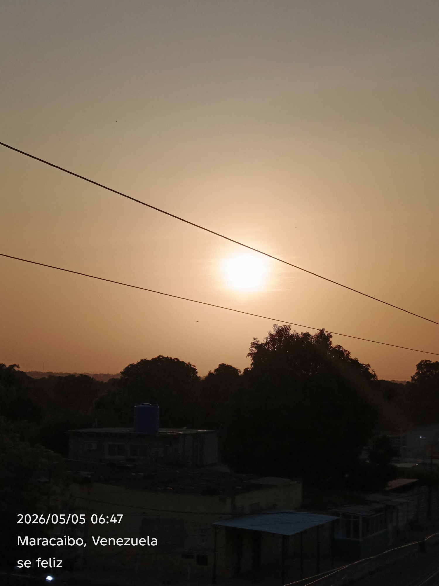 The image depicts a sunrise in Maracaibo, Venezuela, on May 5, 2026, at 06:47. The sun is prominent in the sky, partially obscured by power lines. Below the horizon line, there is a dense collection of trees and the rooftops of buildings. A water tank is visible on top of one of the buildings. The words "se feliz" (be happy) are printed at the bottom of the photo.