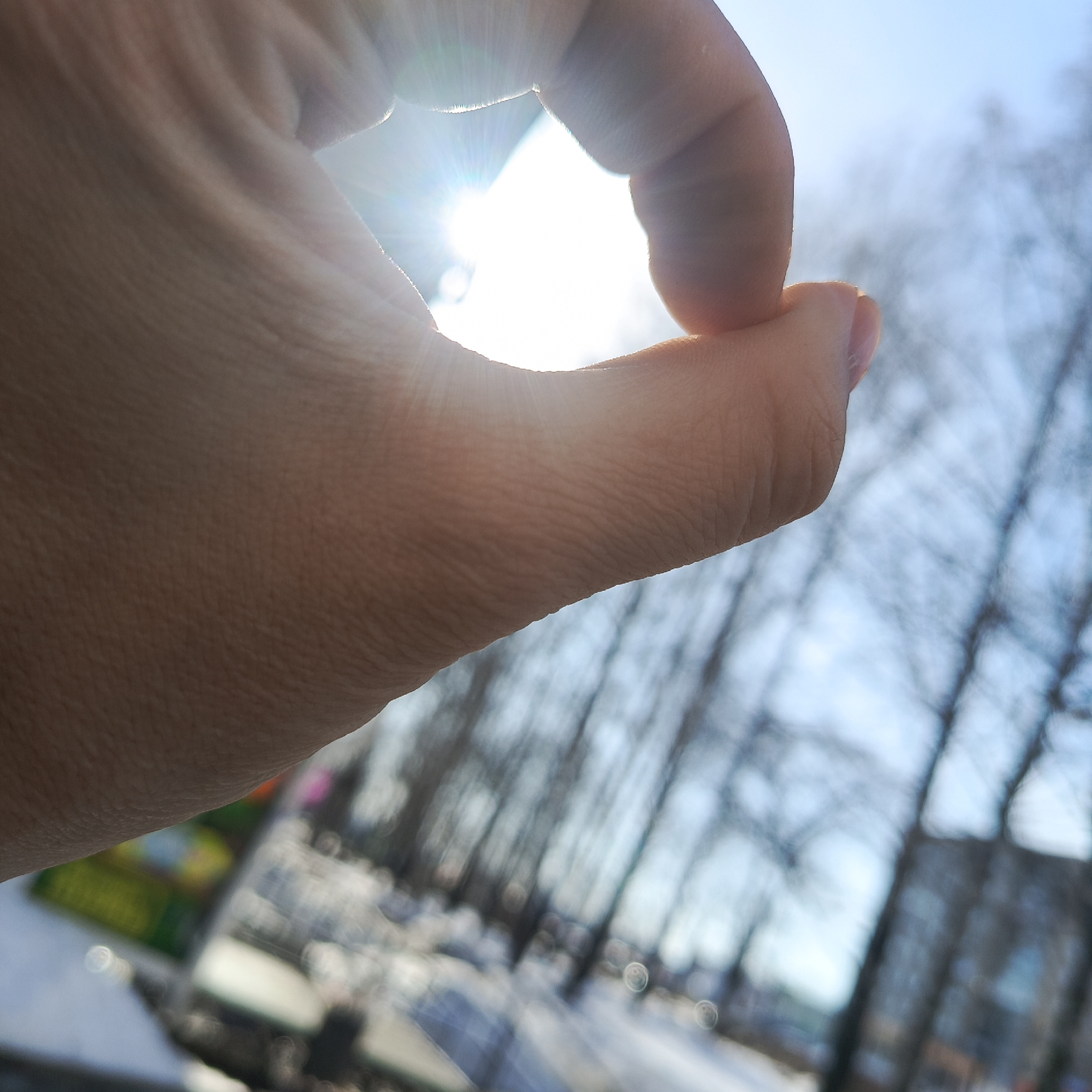 The image shows a person's hand making an "OK" symbol in front of a blurred background. The sun is shining through the circle made by the fingers. The background includes bare trees and a building with a glass facade. There are patches of snow on the ground. The location is potentially in Chaikovsky, Russia. There are no other people visible in the image.