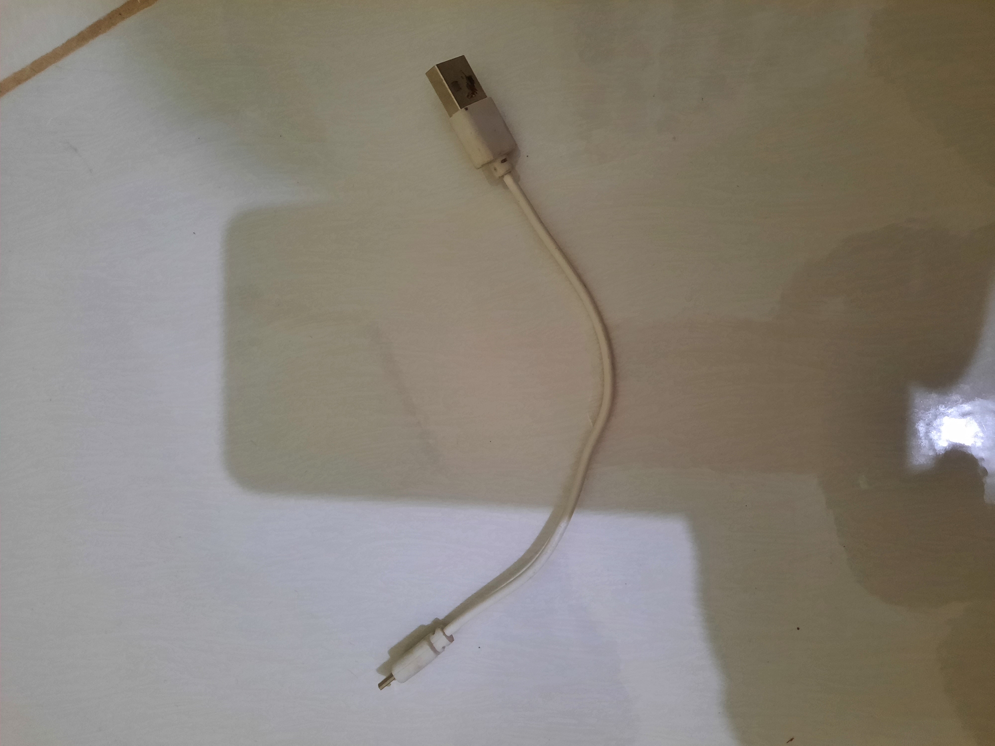 A white USB cable is laid out on a tiled floor. The cable has a micro-USB connector on the left and a standard USB-A connector on the right. The background is a light-colored tiled surface with shadows cast from an unseen light source. The scene is indoors and appears to be a simple, everyday setting.