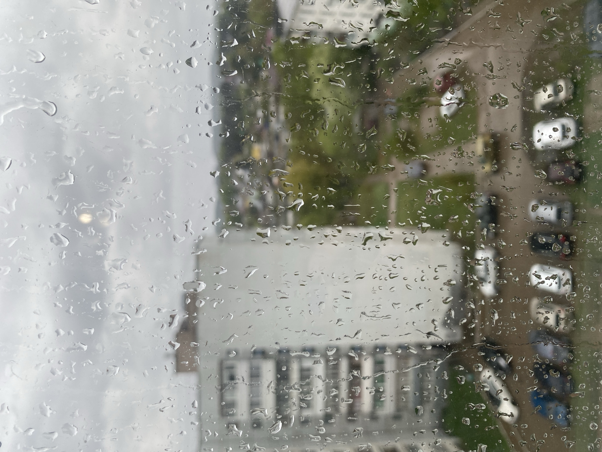 The image shows a view through a rain-covered window. Outside, there is a building with multiple floors and a flat roof. Adjacent to the building is a road with several parked cars. The background includes trees and possibly other buildings, though details are obscured by the rain. The location is likely in a city setting, given the presence of buildings and cars. There is no indication of people in the image. The photograph presents a blurred and distorted view of an urban landscape through the lens of a rain-soaked window. The specific location is the city of Chaikovsky, Russia.