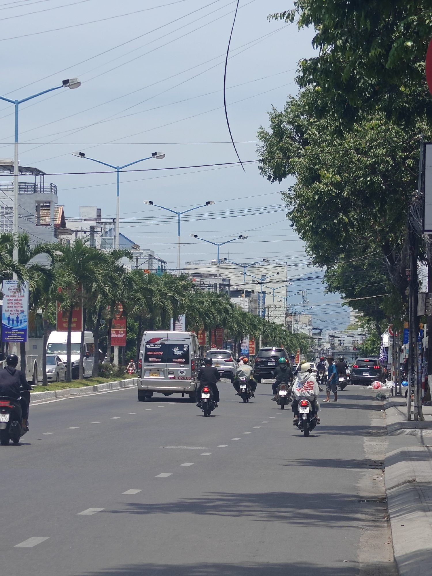 The image depicts a street scene in Bac Nha Trang Ward, Vietnam, on a clear day. The road is paved with asphalt and marked with white dashed lines indicating lanes. On the left side of the road, there is a row of palm trees lining a sidewalk, beyond which are buildings. On the right side of the road, there is a sidewalk with a drainage gutter, and further to the right, a dense canopy of green trees.
Several vehicles are present on the road. In the foreground, two motorcycles are visible, one in the left lane and one in the right lane, both moving away from the camera. The motorcycle on the left is black with a rider wearing a black helmet and jacket. The motorcycle on the right has a rider wearing a white hooded sweatshirt with an American flag graphic and a helmet. Further down the road, there are more motorcycles and cars, including a silver van in the right lane with Vietnamese license plates, a black SUV in the left lane, and a dark colored sedan behind it. Other vehicles are visible in the distance.
Several streetlights with blue poles and white lamps are spaced along the left side of the road. Banners with Vietnamese text are displayed on poles along the sidewalk. Electrical wires are strung overhead. The overall impression is a busy urban street with moderate traffic.