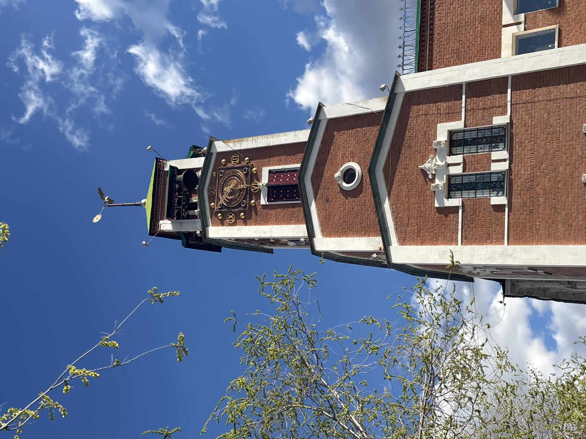 The image captures a view of the Orenburg Puppet Theater in Russia, looking upwards. The building's architecture is characterized by its brick facade and distinct white accents. A prominent clock feature adorns the structure, complete with a weather vane. The sky overhead is a vibrant blue, scattered with white clouds. In the foreground, branches of a tree, displaying emerging green leaves, are visible.