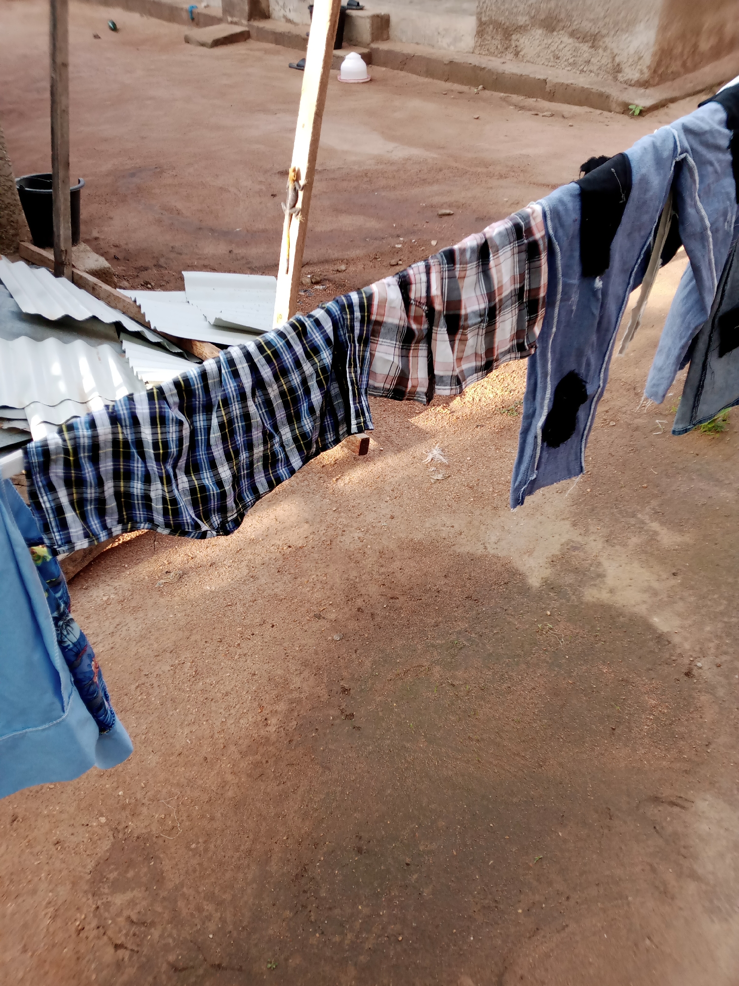 In Jauro Yinu, Nigeria, an outdoor scene shows clothing hanging on a clothesline. The clothesline is suspended between wooden poles. A prominent piece of clothing is a plaid shirt with black, white, blue, and yellow checks, draped across the line. To the right of the plaid shirt, several blue garments with visible black patches hang. On the left, a blue fabric is partially visible. The background consists of a dusty, reddish-brown ground with some scattered debris and a few green sprigs of grass. In the distance, there are structures made of concrete blocks and corrugated metal sheeting, indicating a residential or utilitarian area. A black bucket and a white plastic bowl are also visible in the background, near the wooden supports. Sunlight casts shadows on the ground.