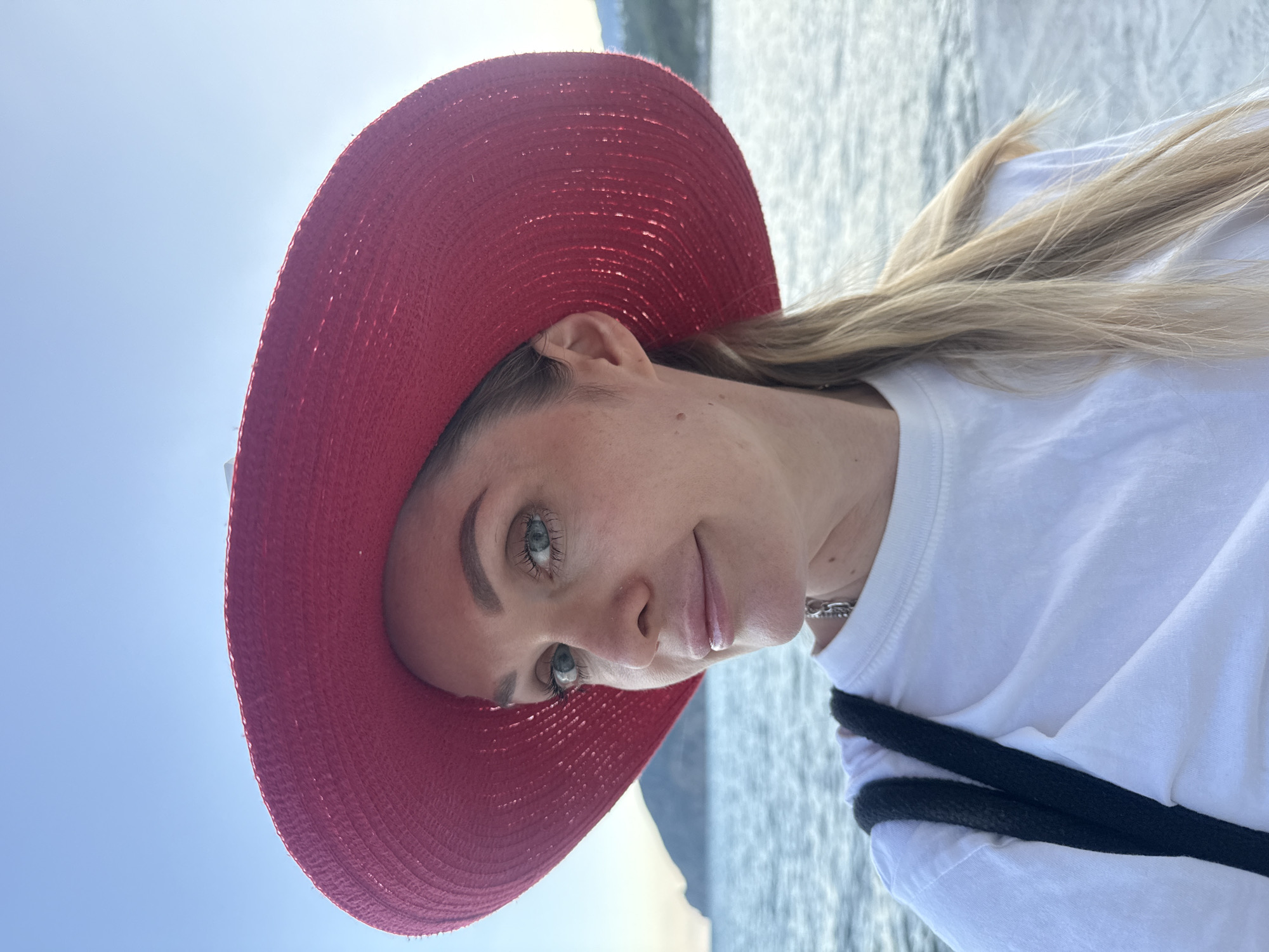 A woman with blonde hair and blue eyes wears a large, red, wide-brimmed hat and a white t-shirt. She is taking a selfie outdoors, with the sea and a distant hill visible in the background under a clear sky. She is wearing a silver chain necklace.