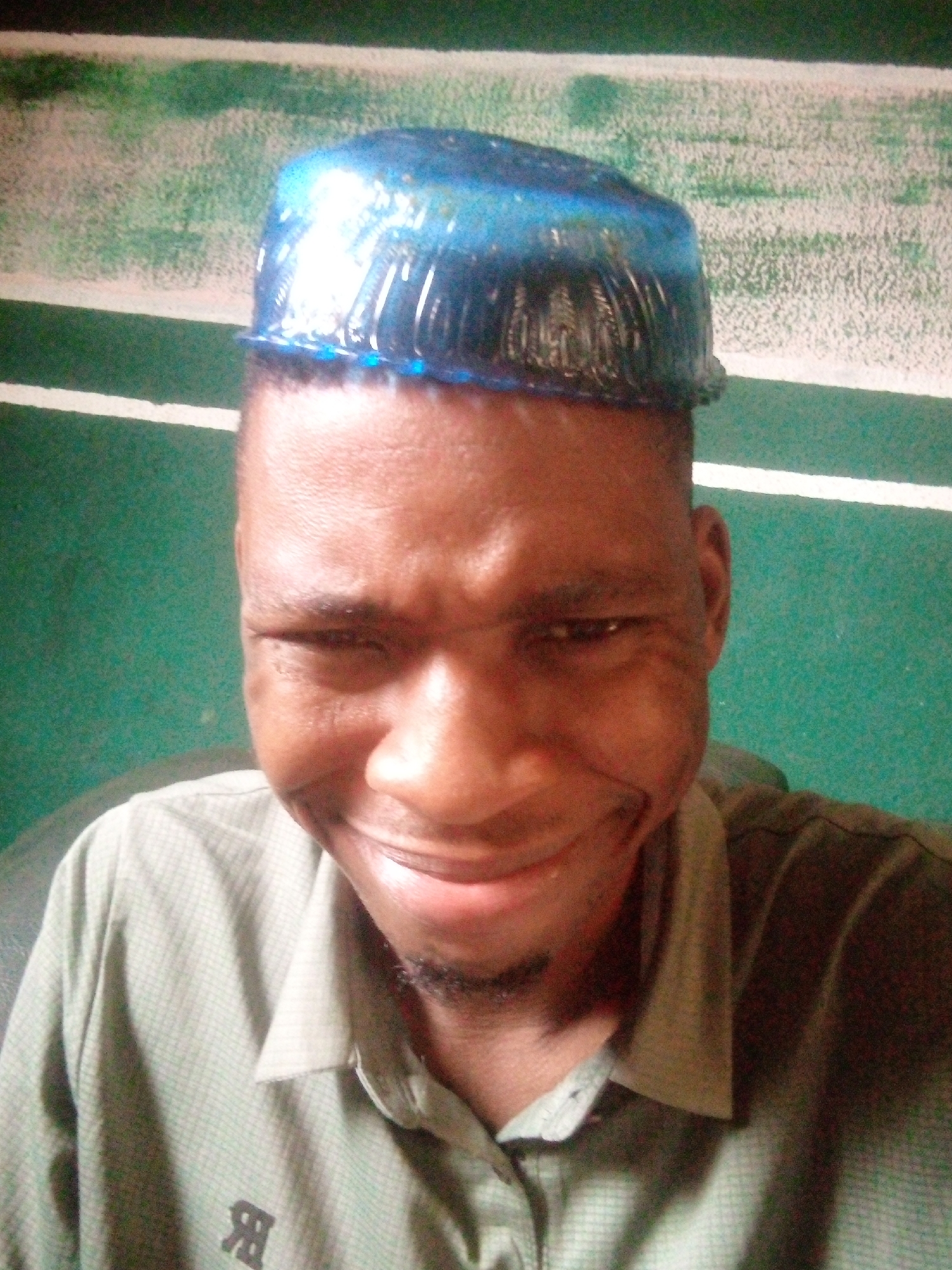 A man is pictured indoors with a blue, plastic container on his head. The background is green with a horizontal white stripe. The man is wearing a button-down shirt that is partially visible. The location is Unknown, Unknown.