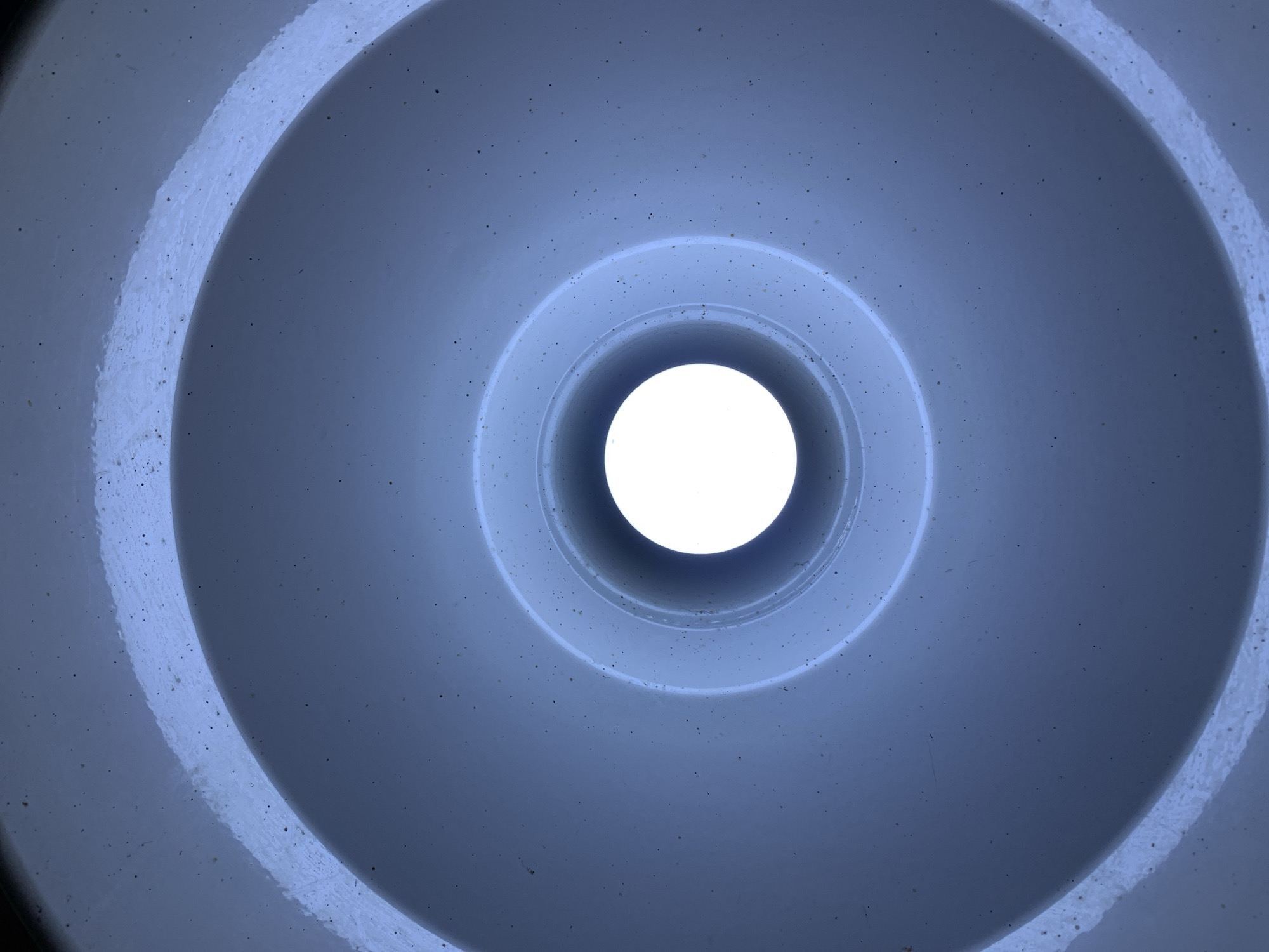 The image depicts the interior of a white, cylindrical structure, possibly a pipe or tunnel, with a bright, circular opening at the far end. The concentric rings suggest a layered or telescoping design. The surface of the structure is speckled with tiny dark spots. The lighting within the structure is predominantly blue-tinged. The location, New Stadium, Nigeria, is irrelevant to the image's content, as the scene is an enclosed space with no visible environmental context. There are no people, actions, or interactions visible in the image.