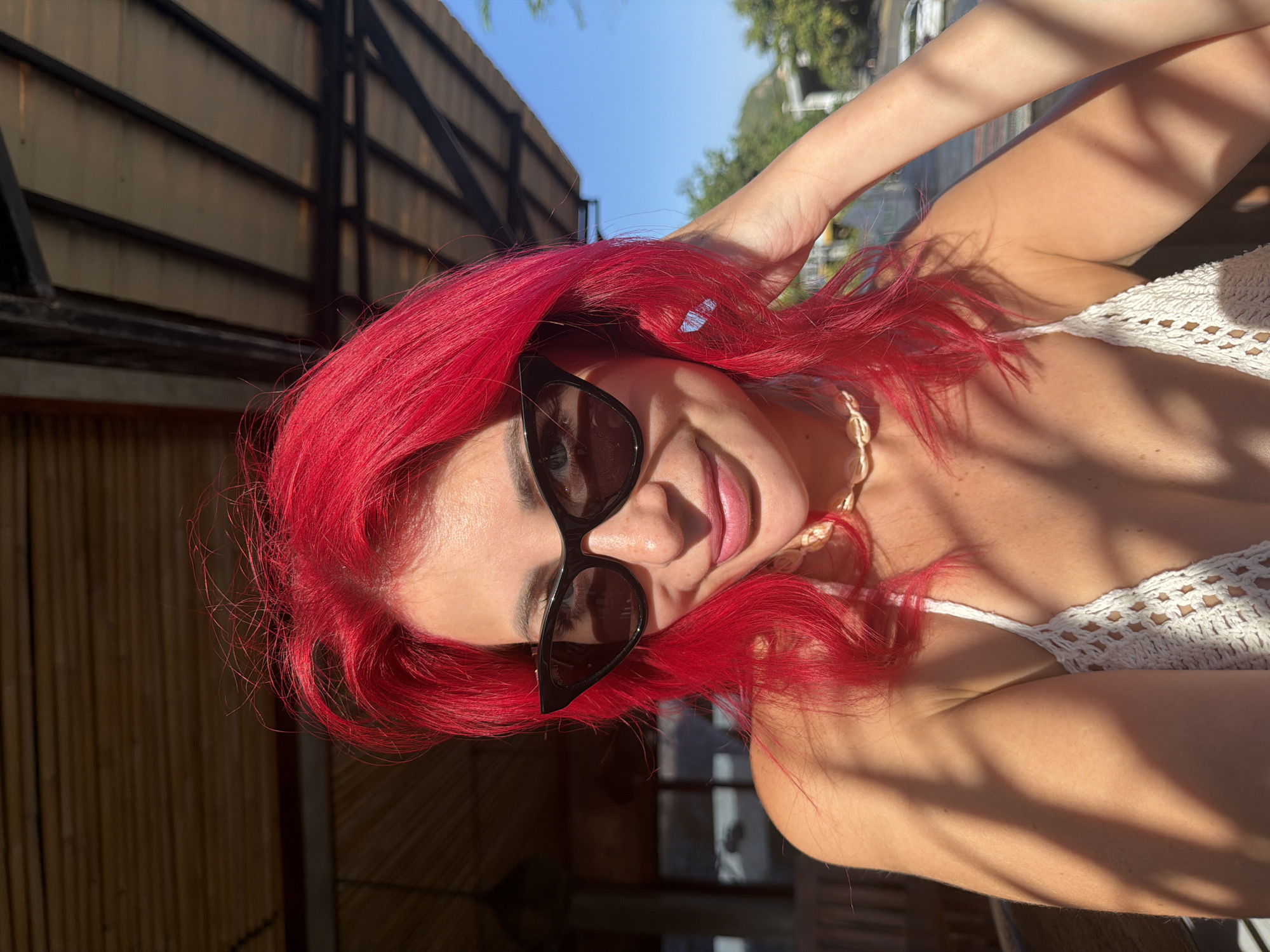 A woman with bright pink hair, wearing black cat-eye sunglasses and a white crochet bikini top, is photographed outdoors in Kute, Indonesia. She is smiling and touching her hair with her right hand. The background shows a thatched roof structure and a clear blue sky. The image is taken from the chest up, with sunlight casting shadows on her shoulder and arm.