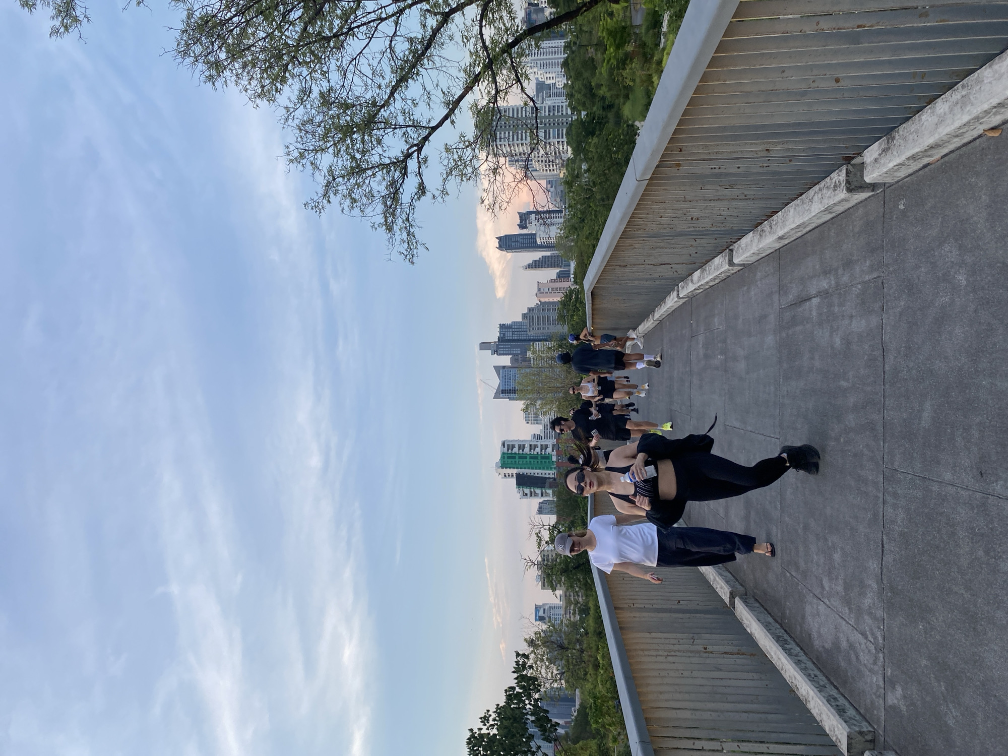 A horizontal, full shot presents an elevated pedestrian walkway in Bangkok, Thailand. The walkway, primarily gray concrete, is slightly curved. A metal railing with horizontal bars is visible on one side. A group of people, roughly ten in number, is walking on the walkway, moving towards the cityscape in the background. The cityscape features a mix of high-rise buildings of varying heights. Vegetation, including trees and bushes, is visible below the walkway. The sky is a gradient of light blue to white, with a few branches of a tree in the upper left corner.
The people on the walkway are wearing casual attire, including athletic wear.