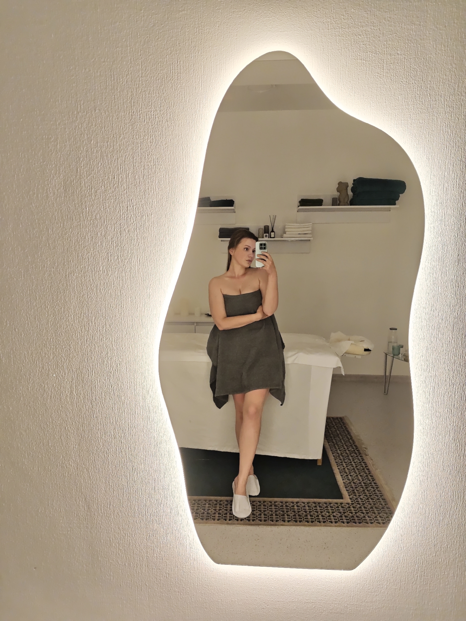 A woman is taking a selfie in a mirror in a brightly lit room. She is wearing a dark gray towel and white slippers. The room appears to be a spa or treatment room, with a massage table and shelves with towels and toiletries in the background. There is a patterned rug on the floor. The mirror has an irregular shape and is surrounded by a light. The setting appears clean and professional. The image does not contain enough information to determine whether it was taken in the city of Vinnytsia, Ukraine.