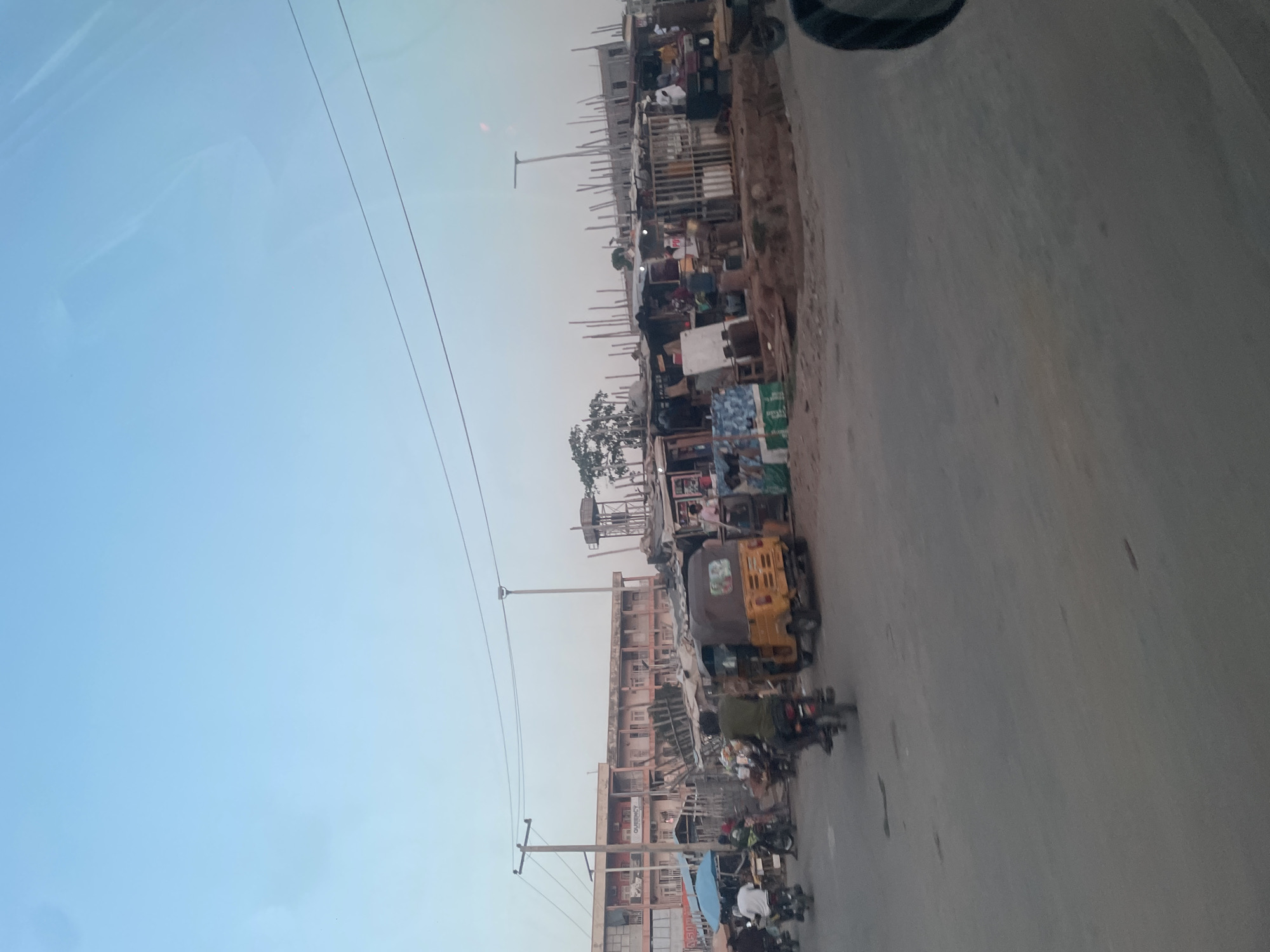 The image depicts a street in Abuja, Nigeria. A road is on the right. Buildings are visible along the left side, some are still under construction with wooden scaffolding. There are power lines in the sky, a mix of building materials and various items are stacked along the side of the road. A yellow tricycle taxi and several motorcycles are on the road. It is daytime. The sky is visible in the background.