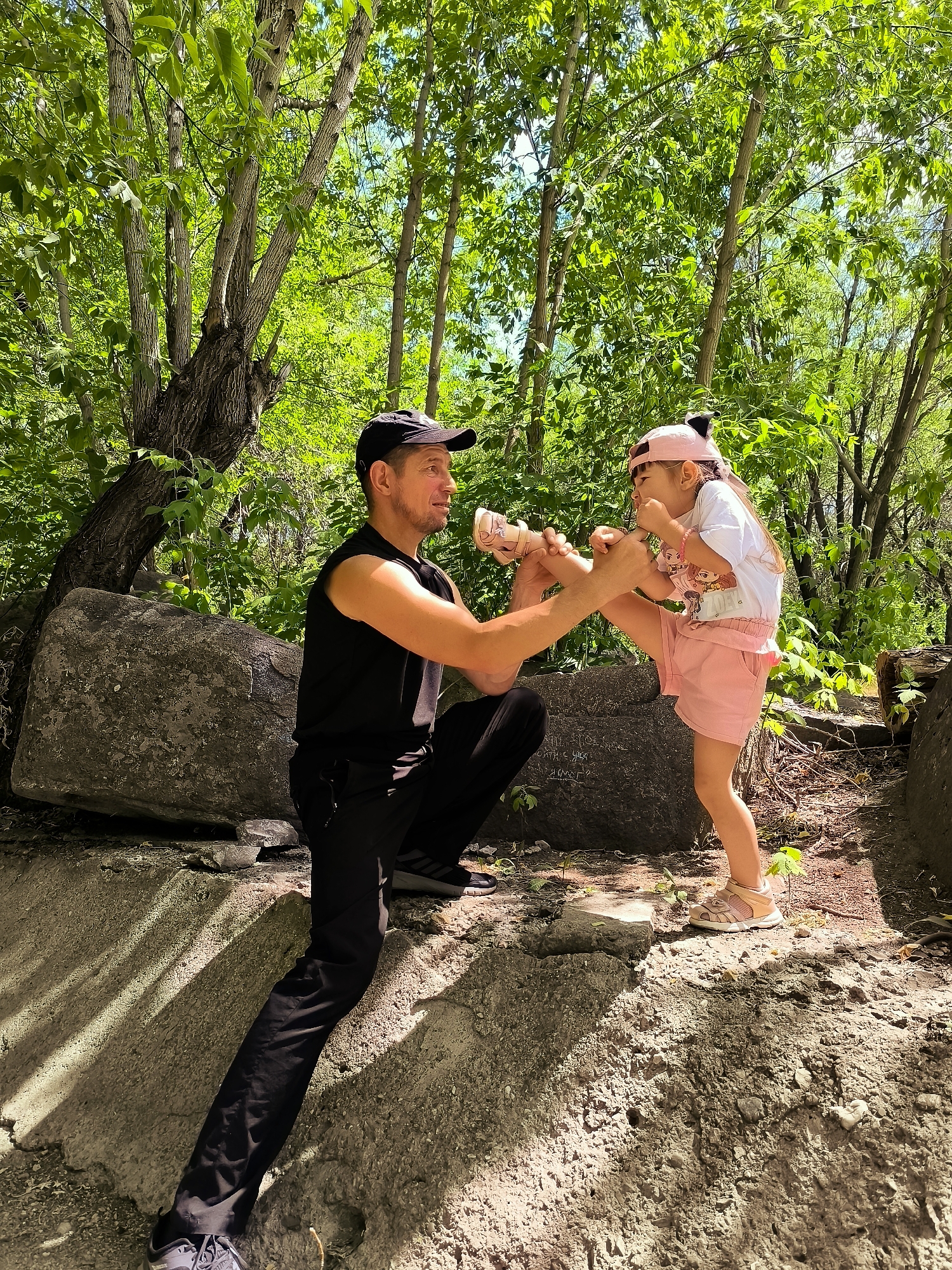 In a sun-dappled forest setting, a man and a young girl are engaged in a playful interaction. The man, wearing a black sleeveless shirt, black pants, and a black baseball cap, is crouched on a rocky incline. He is holding the girl's left leg, which is bent at the knee. The girl, dressed in a pink t-shirt, pink shorts, and a pink baseball cap, stands on the rocky ground, looking at the man and appearing to be laughing or reacting with amusement. She has on light pink sandals.
The environment is lush and green, with mature trees and dense foliage suggesting a natural outdoor location. Large rocks are scattered throughout the scene, forming part of the terrain. The sunlight filtering through the trees creates dappled patterns on the ground and the subjects. The overall atmosphere suggests a lighthearted moment between the two individuals.
There is no visible text in the image. The scene appears to be taking place during the daytime, under clear skies, given the brightness and the shadows.