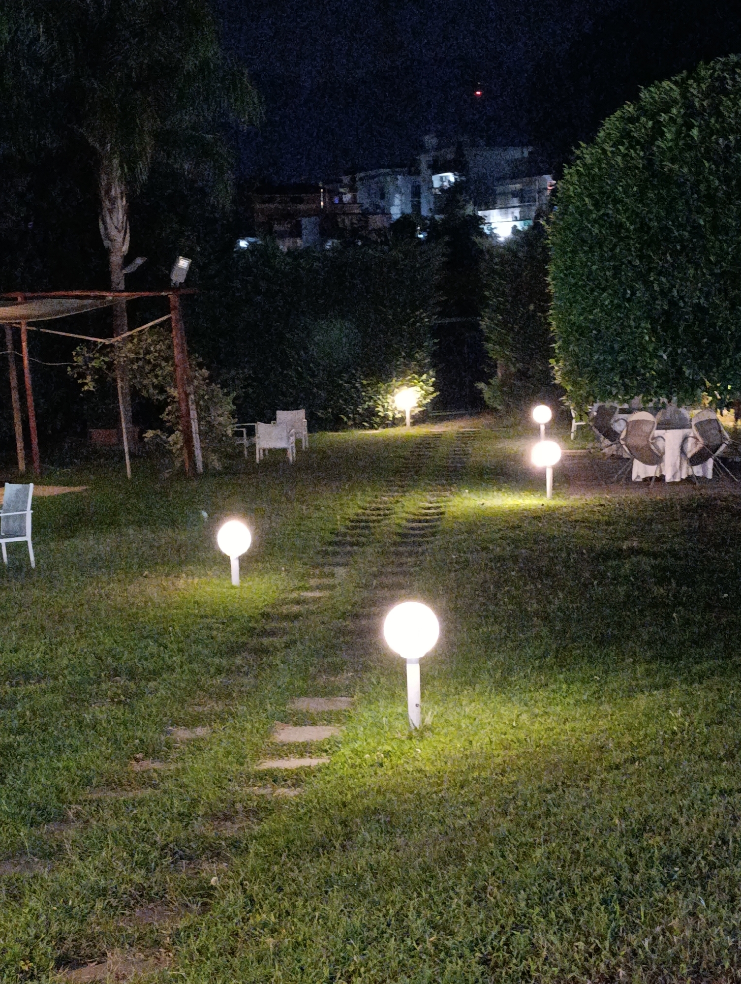This is a nighttime photograph of a garden area in Castellammare di Stabia, Italy. The scene features a pathway of stone pavers winding through a well-maintained lawn, illuminated by several spherical garden lights. To the left, a white plastic chair is visible. Further into the garden, a rustic wooden pergola structure stands with some sort of dark-colored covering. In the background, beyond a dense hedge, buildings are dimly lit. On the right side of the image, a round table is set with white tablecloths and chairs, suggesting an outdoor dining area. A large, dark green bush dominates the right edge of the frame. The overall impression is that of an outdoor event space or a restaurant's garden at night, decorated with landscape lighting.