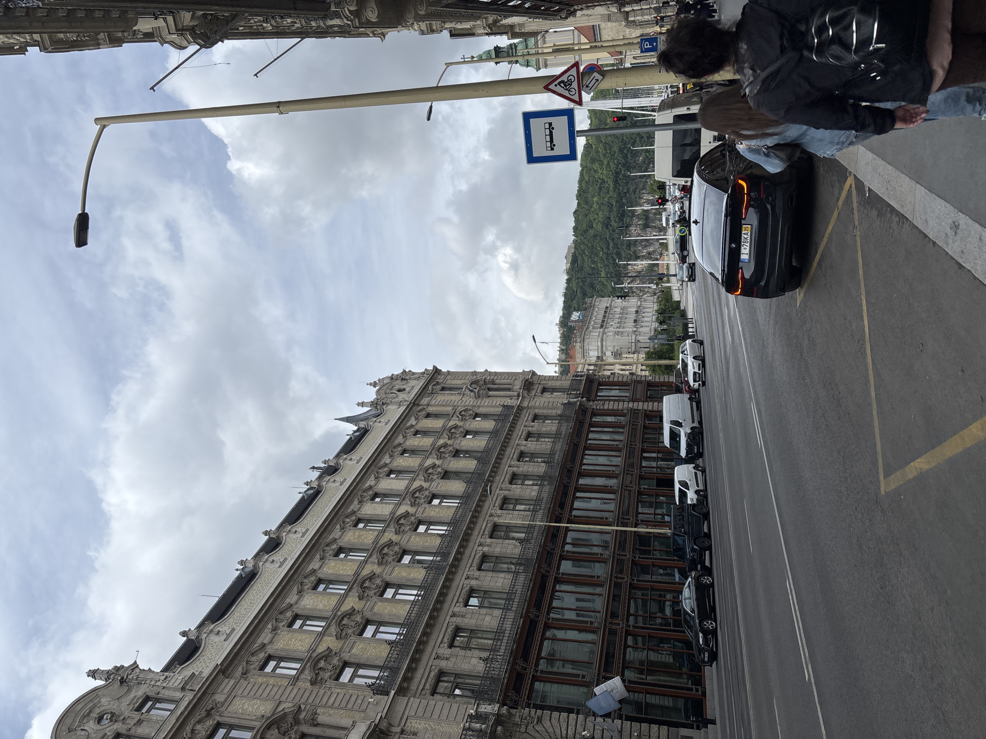 This image shows a street scene in Budapest, Hungary. The dominant feature is a tall, ornate building on the left side of the frame. Across the street from the building is a row of parked cars, including a black BMW with a license plate that reads "I-78KA25". A couple is standing on the sidewalk near the BMW. Above the street, there are street signs, including a bus stop sign and a sign indicating a bicycle path. In the background, there is a hillside with buildings on it. The sky is cloudy.