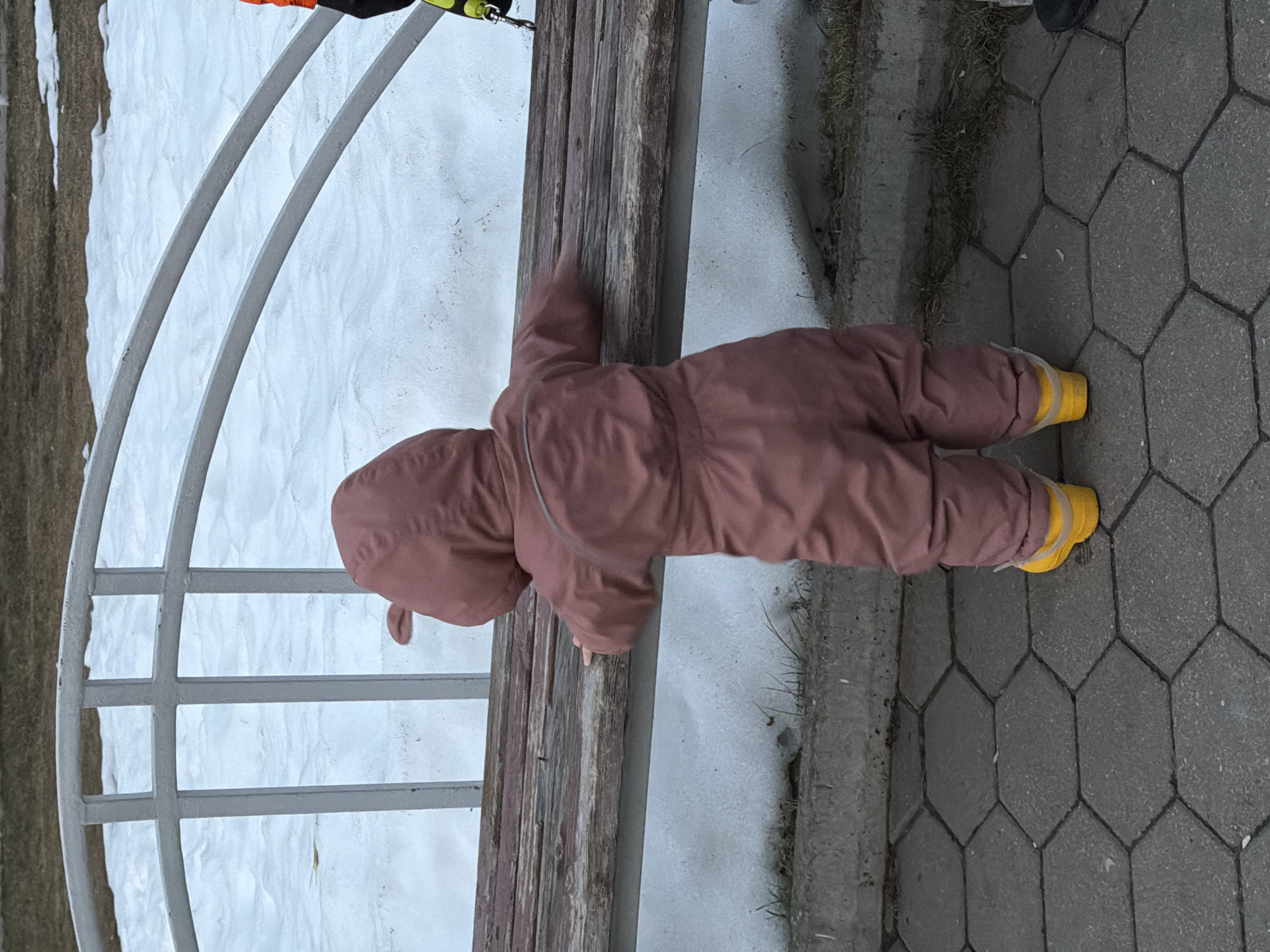 A small child in a reddish-brown snowsuit and yellow boots is crawling on a stone-paved surface near a wooden bench with a white metal frame. The bench is located next to a patch of snow. The location appears to be outdoors, likely a park or recreational area in a city that experiences cold weather, which could potentially be Chaikovsky, Russia.
