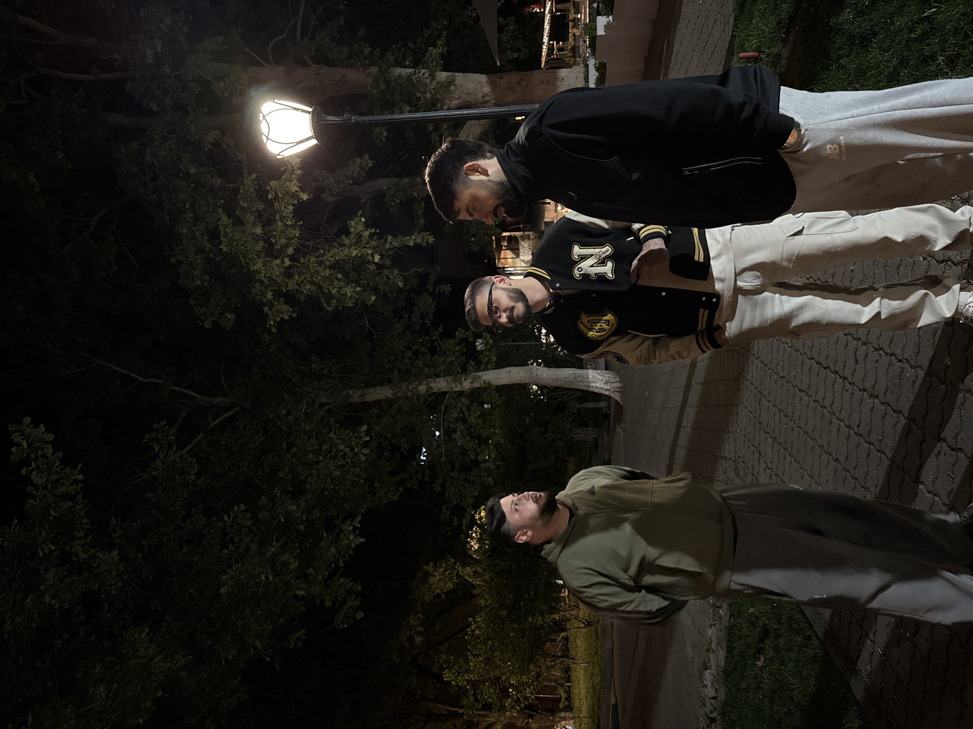 Three men are standing on a paved area that appears to be a sidewalk in a park or plaza. The lighting indicates it is nighttime. The area is illuminated by a classic-style lamppost. The environment includes trees and vegetation, suggestive of a park setting. The men are dressed in casual clothes. The man nearest the camera is in an olive-green hoodie and dark pants. The middle man is wearing a letterman jacket with the letter "N" on it, and light-colored trousers. The farthest man is wearing a black jacket and gray sweatpants. They appear to be in conversation or casually looking at something together.
Based on the architectural style of the lighting and the paving, and given the information provided, it is very likely that this photo was taken in the city of Yerevan, Armenia.