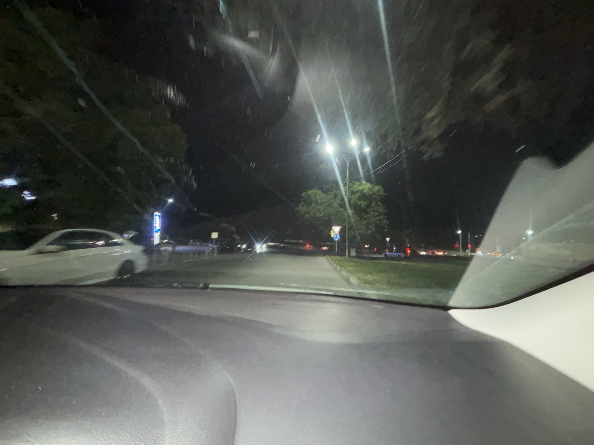 The image is taken from inside a vehicle at night, looking out through the windshield. The scene appears to be a street in Grozny, Russia, characterized by streetlights illuminating a road lined with trees. On the left side of the road, a white car is parked. Further down the road, there are other vehicles and streetlights. The road itself is paved and appears to be clear. The overall impression is of a nighttime drive through a city environment.