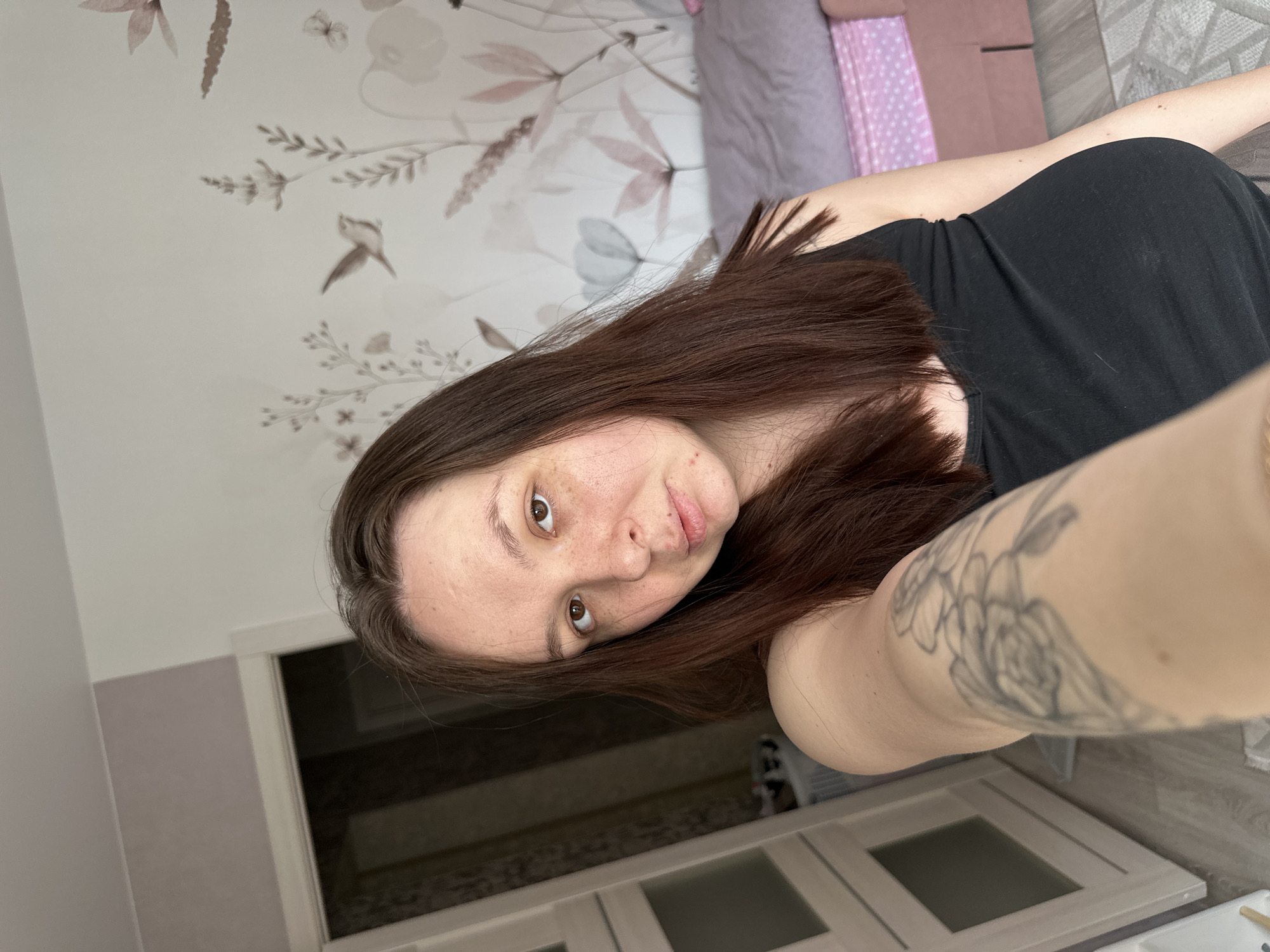 The image displays an adult female in a residential interior located in Chaikovsky, Russia. The subject, centered in the frame, has long dark brown hair, fair skin with noticeable freckles, and brown eyes, looking directly at the camera with a neutral expression. She is wearing a black tank top, and a multi-floral tattoo is visible on her extended left forearm, which holds the camera for a self-portrait. To the left of the subject, a white-framed doorway with a partially open light-colored door leads into a darker room exhibiting vertical striped patterns. To the right and behind the subject, a wall is covered with light-toned wallpaper featuring a pattern of birds, butterflies, and various plants in muted pink, grey, and brown hues. A bed or sofa, partially visible on the far right, is covered with a light purple-grey blanket and a pink patterned sheet or upholstery underneath. A light-colored, patterned rug is visible on the floor in the lower right corner.