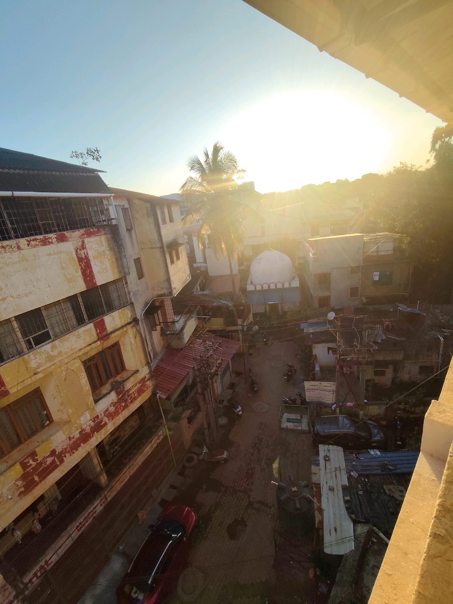 This is a high-angle, outdoor, urban scene taken during daylight, likely in Belagavi, India. The sun is setting or rising, casting a strong golden light and lens flare across the image, obscuring details in the background. In the foreground, a weathered, multi-story building with yellow and red paint is visible on the left. Its facade shows signs of age and disrepair, with barred windows. The building appears to be residential or mixed-use.
Below the building, a narrow street or alleyway is visible, lined with smaller structures, some with corrugated metal roofs. A mosque with a white dome is present further down the street. Several motorcycles and a dark-colored car are parked on the street. The environment appears dense and somewhat cluttered, typical of a bustling urban area. A palm tree is visible in the mid-ground, illuminated by the sunlight. On the far right of the image, a concrete ledge or balcony edge is visible, suggesting the photograph was taken from an elevated position, possibly another building or balcony. The overall impression is one of a lived-in, working-class urban neighborhood bathed in the warm glow of the sun.