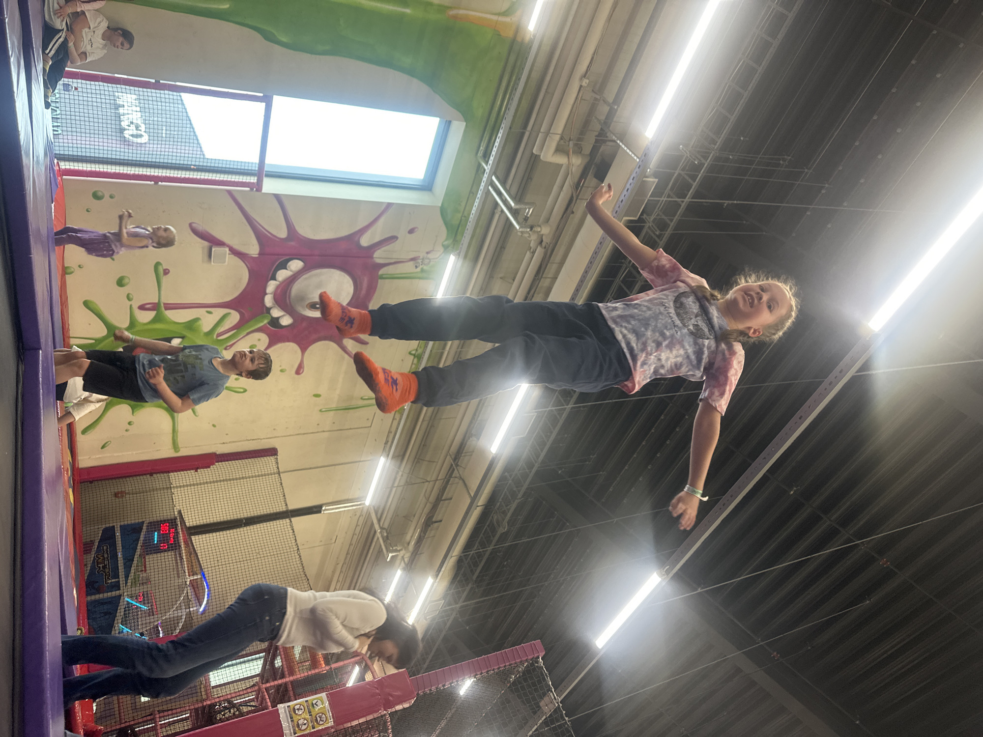 The image shows the interior of a trampoline park in Riga, Latvia. A girl is mid-air, seemingly jumping on a trampoline, wearing a tie-dye shirt, dark pants, and orange socks. Other people are visible in the background, including two adults and three children. The walls of the trampoline park are decorated with colorful cartoonish designs. The ceiling shows the building's infrastructure with visible pipes and lighting fixtures. Natural light enters through a window. Trampolines are seen within the park.