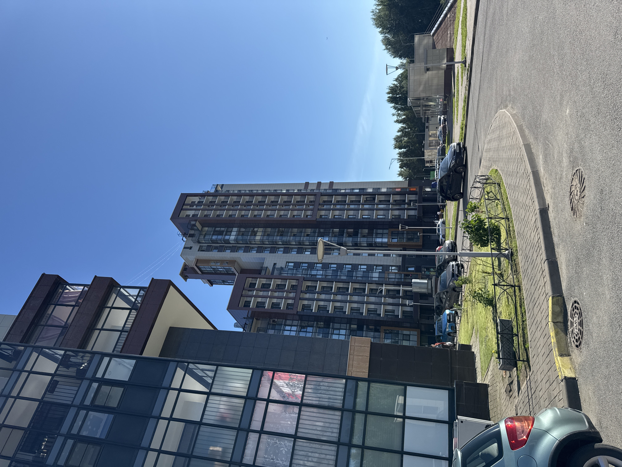 The image depicts a bright, sunny day in a modern residential complex in Saint Petersburg, Russia. The foreground features a paved road and sidewalk with a sewer grate. To the left, a blue-green car is parked. Further into the scene, a dark grey building with large glass windows and balconies dominates the left side of the frame. Adjacent to it, and slightly further back, are two tall apartment buildings with a multi-colored facade, featuring numerous windows and balconies. A grassy area with a decorative fence separates the buildings from the road. Several cars are parked in the vicinity of the buildings. In the background, a line of green trees forms a natural barrier, with a clear blue sky overhead. The architecture suggests a recently constructed urban development.