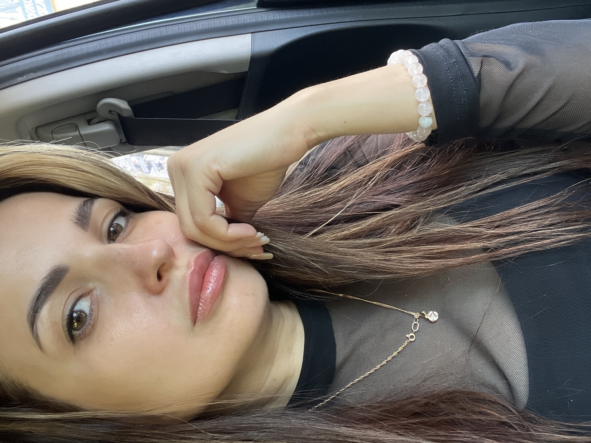 A woman is in a car. She has brown hair, and is wearing a black mesh top with a necklace and a bracelet. She is holding her face with her hand.