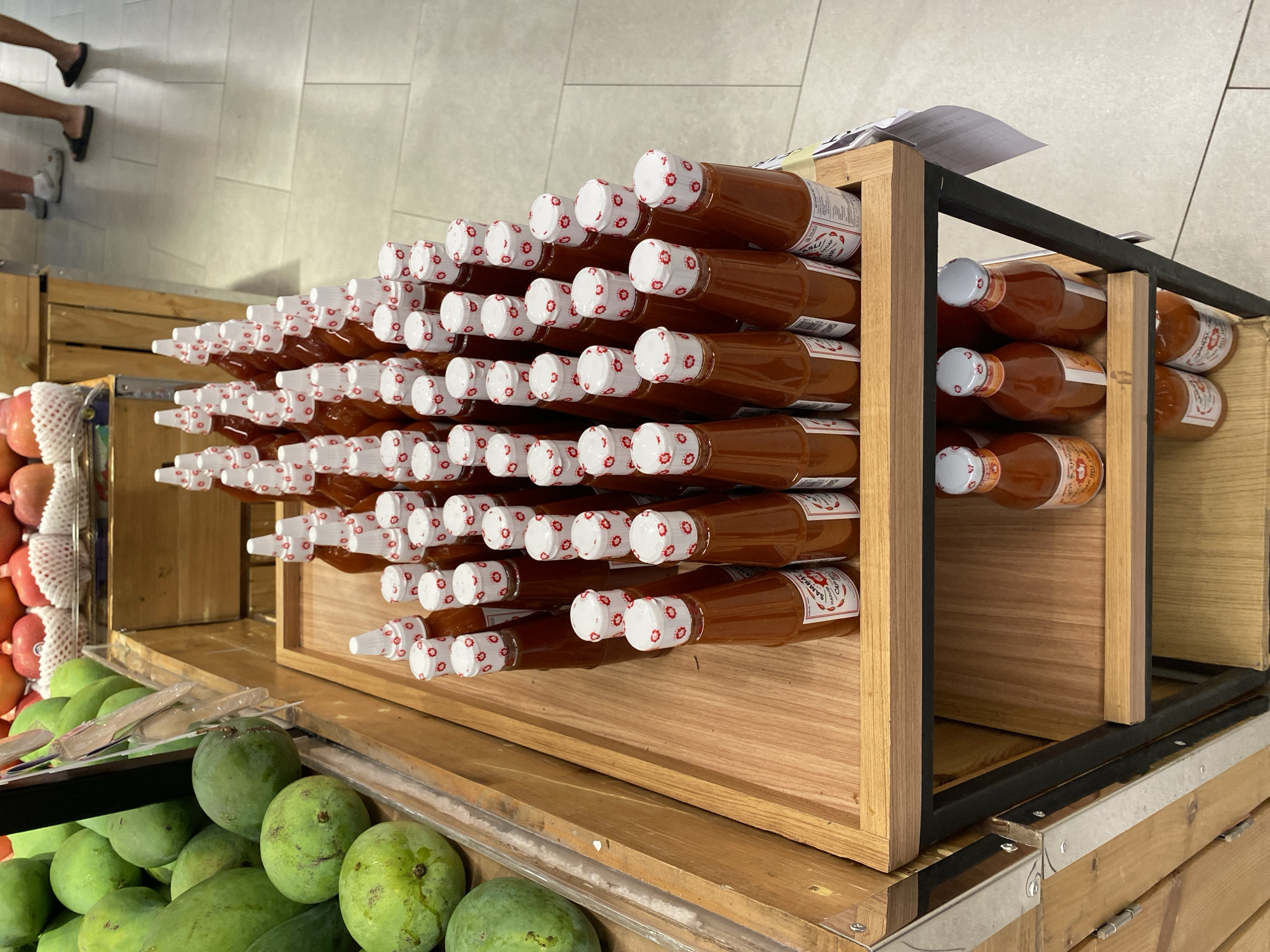 The image depicts a retail display of bottled sauces, likely sambal, in a supermarket in East Jakarta, Indonesia. The sauces are arranged in several rows on wooden shelves. The bottles are amber glass with white caps adorned with a red floral pattern. Some of the labels are partially visible, suggesting they are local brands. In the background, a display of green mangoes is visible to the left, and the legs of shoppers are visible to the right, indicating the setting is a public market or grocery store. The floor is tiled in a light gray color. The overall scene is a snapshot of a product display within a bustling commercial environment.