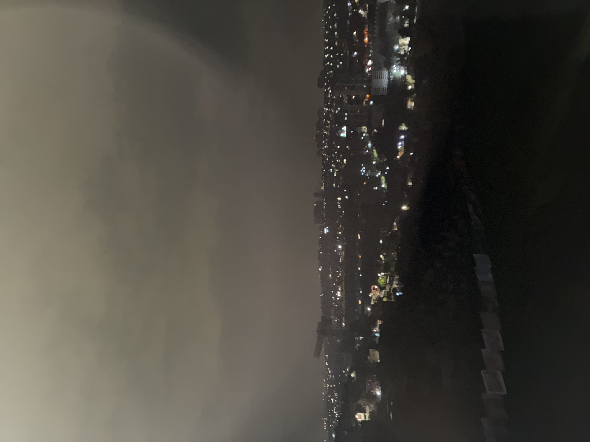 The image shows a nighttime view of a city, possibly in Huixquilucan de Degollado, Mexico. The left side of the image is dominated by dark, overcast sky. The city itself is characterized by scattered lights from buildings and streetlights. The right side of the image fades into complete darkness. No people are visible.