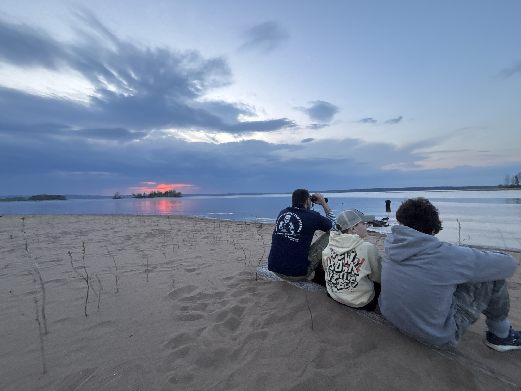 Three individuals sit on a sandy beach at Vekoshinka, Russia, watching a sunset over a body of water with islands. The first person on the left is an adult male using binoculars. He is wearing a dark-colored t-shirt with an image on the back. The two people to the right are younger. One is wearing a hoodie and a baseball cap, the other one is wearing a hoodie. The ground is sand with sparse vegetation. The sky is cloudy, with the setting sun reflecting on the water.
