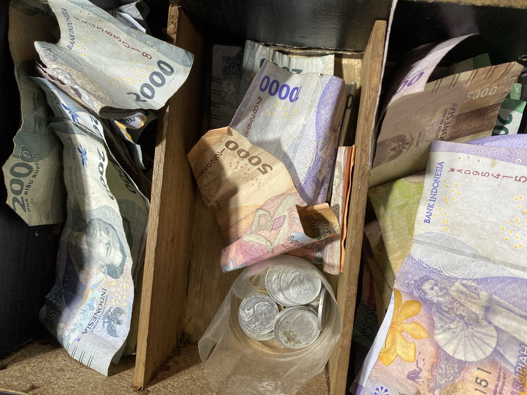 The image depicts a close-up, top-down view of a wooden box filled with Indonesian currency, both banknotes and coins. The banknotes are of various denominations, including 2000 and 5000 Rupiah, and some appear to be newer issues with holographic elements. The coins are contained within a clear plastic bag. The lighting suggests an indoor setting, possibly a drawer or a small safe. The image focuses on the contents of the box, with no discernible background elements that would indicate a specific location within Kasihan, Indonesia.
