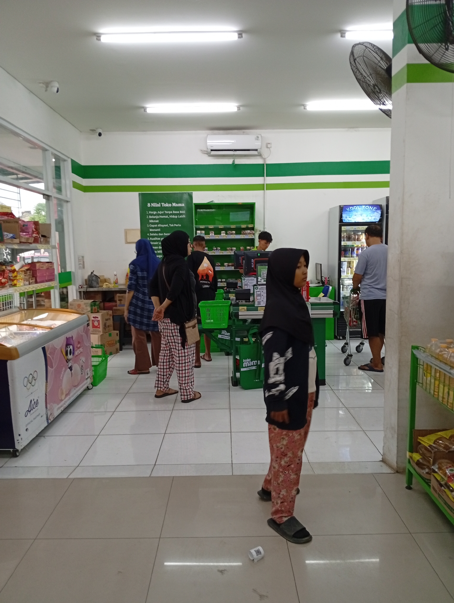 The image depicts the interior of a minimarket, possibly located in Babakan, Indonesia, characterized by its clean, well-lit environment and green and white decor. In the foreground, a young person wearing a black hijab and patterned trousers stands facing left, with their right hand resting on their hip and their left hand visible. To the left, an open freezer with a colorful advertisement featuring cartoon characters is visible, labeled "Alice". Behind the freezer, shelves are stocked with various products.
Further into the minimarket, several other individuals are present. A person in a blue and white checkered hijab stands near a checkout counter, facing away from the camera. Another person, wearing a black t-shirt with an Indonesian flag emblem on the back, is seen from behind, walking towards the right. A man in a light blue t-shirt and dark shorts is seen from the side, pushing a shopping cart towards a refrigerated display case. The display case has a sign at the top that reads "KOOL ZONE".
The environment includes fluorescent lighting, air conditioning units mounted on the wall, and multiple security cameras positioned on the ceiling. Several green shopping baskets are scattered throughout the store, and a large green sign with white text, listing "8 Nilai Toko Mama", is prominently displayed on the wall. The floor is tiled with large, light-colored square tiles. On the right side of the image, a green shelving unit displays various food items. A small white receipt with a QR code is on the floor in the lower right portion of the image.