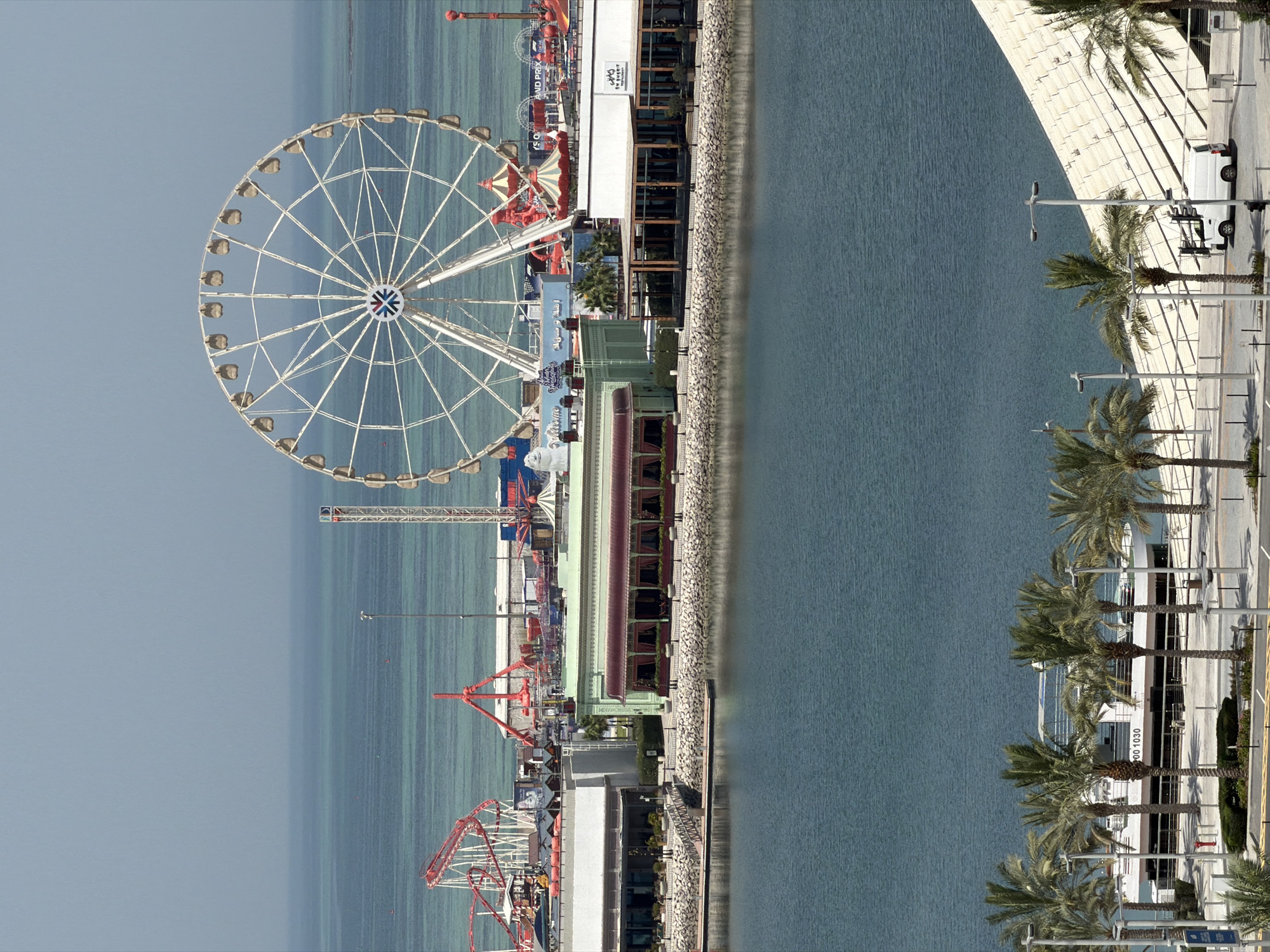 An aerial, medium shot captures an amusement park situated on a waterfront. Dominating the frame is a large white Ferris wheel, its circular structure and spokes clearly defined against the blue sky. To the left of the Ferris wheel, a red roller coaster with its distinctive track design is visible, and various other amusement park rides and structures are scattered throughout the scene, some painted in vibrant colors. In the foreground, a calm body of water stretches across the width of the image, reflecting the light. Beyond the park and across the water, a thin strip of land is visible, with a hazy horizon line separating the sea from the sky. Several palm trees are planted along the waterfront promenade in front of the amusement park buildings, which appear to be low-rise structures with large windows and outdoor seating areas. A white boat with the number "00 1030" visible on its side is docked near the shore. A white van is parked on the paved area in the lower right corner of the frame. The overall scene suggests a recreational area designed for entertainment and leisure.