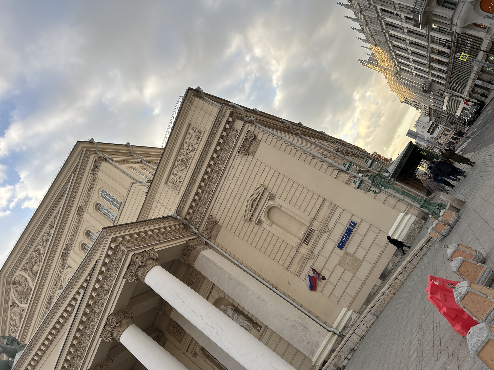 This medium-angle photograph captures a bustling city street scene dominated by two distinct architectural styles under an overcast sky. On the left, a grand Neoclassical building with a light-colored facade features large white columns with ornate capitals, intricate friezes, and arched windows. A blue street sign on its wall clearly reads "Театральная пл." (Teatralnaya pl., meaning Theatre Square) and the number "1," identifying the location as Moscow, Russia, specifically the Bolshoi Theatre. Faintly etched below the street sign are the letters "CCCP." A Russian flag and another red, blue, and white flag (likely Serbian) are mounted on the building.
On the right side of the frame, across the street, stands a tall, intricately designed building with a darker facade, characterized by numerous windows and distinctive pointed spires along its roofline, suggesting a Gothic Revival architectural style. This building is illuminated by soft, low-angle light, possibly from a setting or rising sun, contrasting with the mostly cloudy sky.
Several pedestrians are visible on the paved sidewalk. One person in a black coat walks past the Neoclassical building, while a small group is gathered further down the street near a green canopy. In the immediate foreground, red plastic road barriers and weathered stone-like bollards line the edge of the sidewalk. A green, ornate lamppost stands near the street sign. The scene captures a blend of historical architecture and daily urban life.