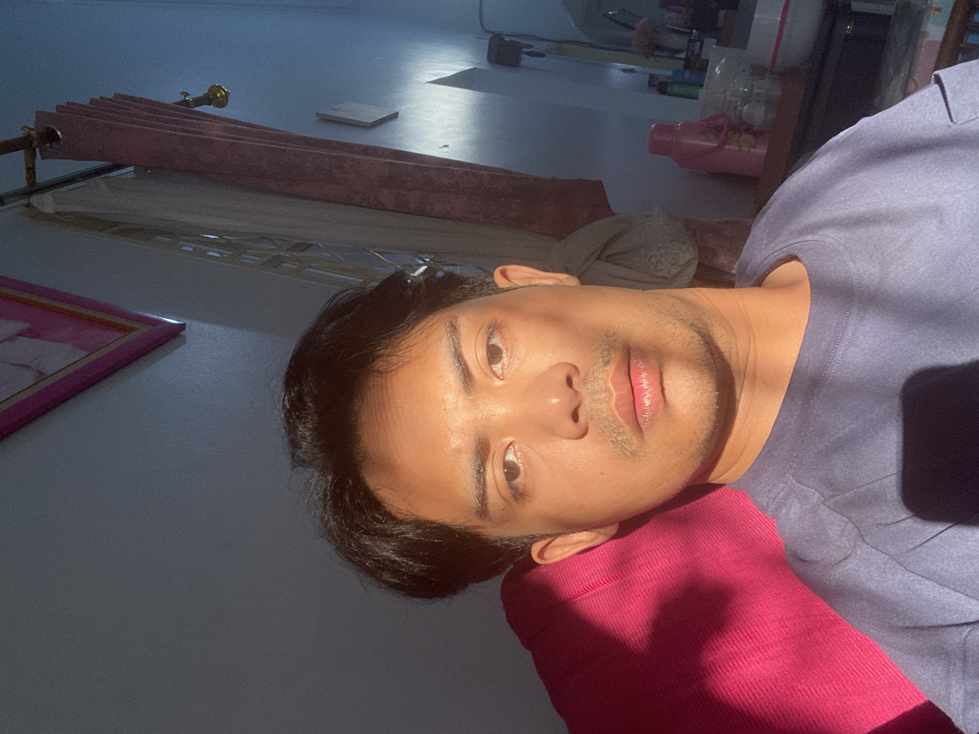 The image shows a close-up, selfie-style shot of a person of Southeast Asian descent, likely male, sitting indoors. The person is positioned in the left half of the frame, facing slightly to the right. A bright light source, possibly sunlight, illuminates the right side of their face, creating a strong contrast with the shadowed left side. They have dark hair, prominent eyebrows, and appear to be wearing a dark blue or gray t-shirt. Behind the person, on the left, is a bright pink textured surface, possibly a cushion or piece of furniture. To the right, the background shows a wall with a pink-toned curtain or drape, a window with a lattice design, and various objects on a shelf or surface, including a pink bottle. The overall lighting suggests it is daytime, with strong directional light. The location of Dukuh, Indonesia, is not visually ascertainable from the image itself.