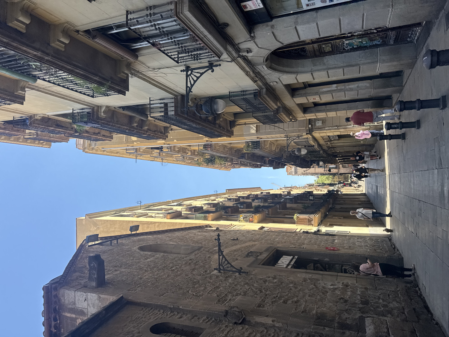 This media file captures a narrow, historic street or alleyway in Barcelona, Spain, with tall buildings rising on both sides towards a clear blue sky. The architecture features multi-story structures made of light-colored stone and stucco, adorned with numerous balconies that display ornate wrought-iron railings, some containing small planters with greenery.
The street is paved with flagstones or large tiles, and a line of black bollards runs along the right side of the walkway. The bright, overhead sunlight suggests it is midday or early afternoon on a clear, sunny day, casting distinct shadows.
Several pedestrians are present. In the foreground on the right, a person wearing a light pink top and dark trousers stands near a building. Further back on the right, another individual in a white t-shirt and dark pants is visible. In the mid-ground, two people walk side-by-side, one in a red shirt and light pants, the other in a light pink top. More figures are visible walking further down the street, indicating everyday pedestrian activity.
A prominent, older building made of darker, rough-hewn stone occupies the lower left and extends into the mid-ground, possibly an ancient structure or part of a historic church. An ornate, black metal street lamp fixture is attached to one of the building facades. While some signs and potential text are present on various buildings and shop fronts, they are too distant or indistinct to be clearly legible in the image.
