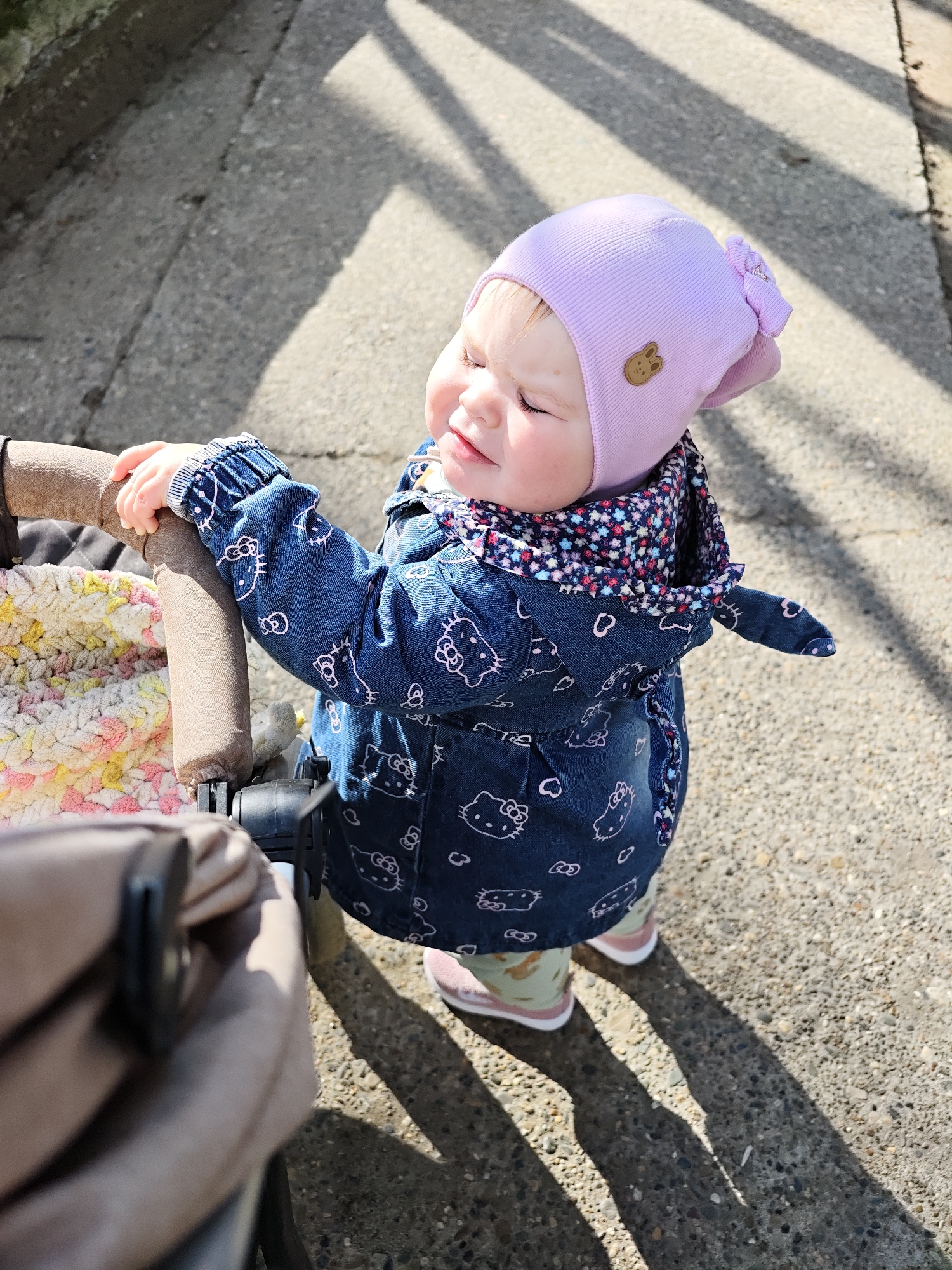 A toddler stands next to a stroller on a concrete surface. The toddler wears a light purple hat, a blue jacket with a "Hello Kitty" print, and pink and green shoes. The stroller contains a multi-colored blanket. The background suggests an outdoor urban setting with shadows cast on the ground. The location is the city of Chaikovsky, Russia.