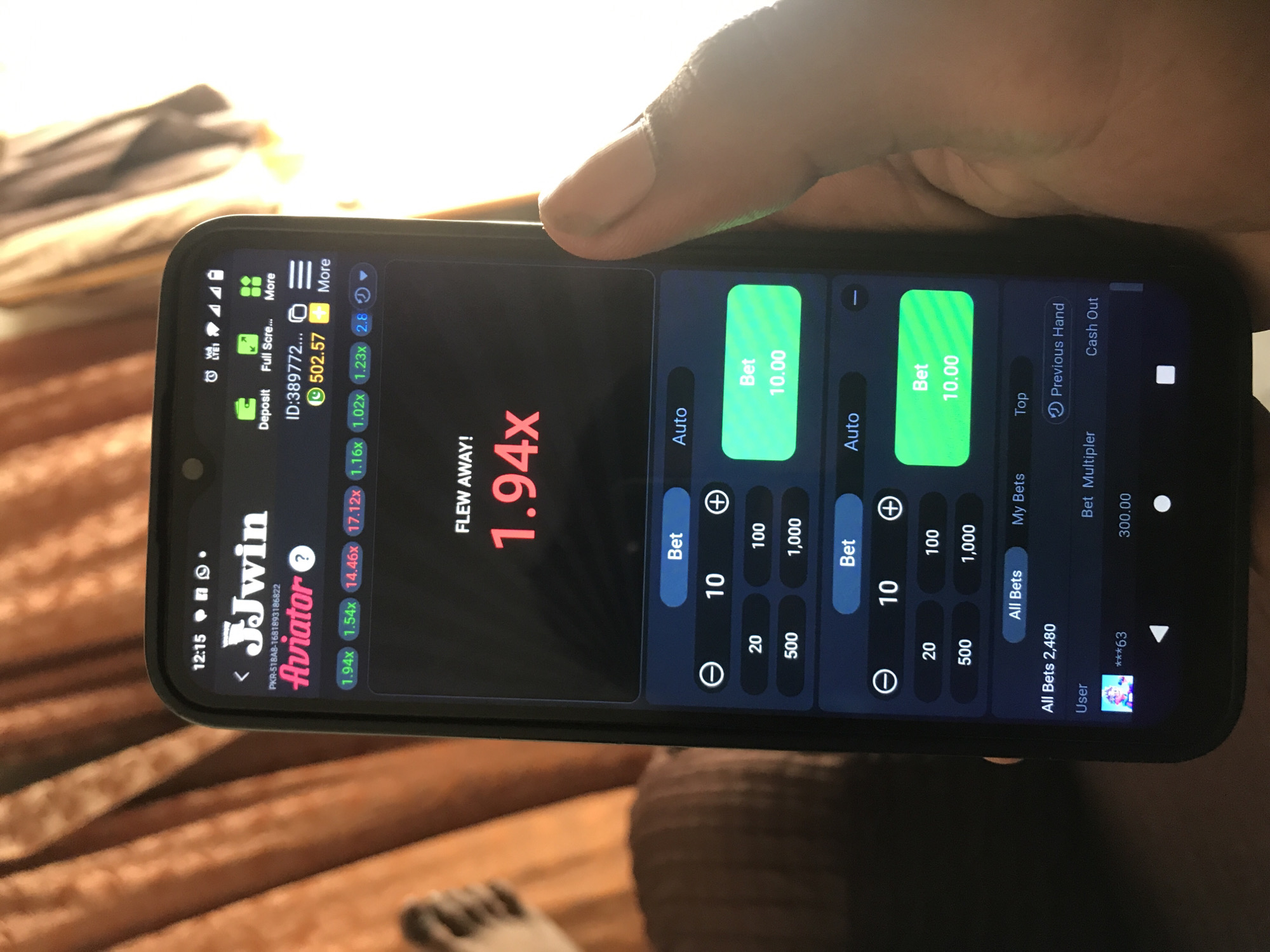 A person is holding a black smartphone in portrait orientation, displaying an online gambling or gaming application called "JJwin Aviator." The game interface is dark-themed with green and blue accents. The screen shows the current game status, "FLEW AWAY! 1.94x", indicating a multiplier achieved in the game. Below this, a series of past multipliers are visible: 1.94x, 1.54x, 14.46x, 17.12x, 1.16x, 1.02x, 1.23x, and 2.8x.
The user's account balance, "502.57", is displayed near the top, along with options for "Deposit" and "Full Screen." The phone's status bar shows the time as "12:15," along with LTE1 network strength, Wi-Fi, and battery icons.
The lower part of the screen features two identical betting sections. Each section has "Auto" toggles and large green buttons labeled "Bet 10.00", indicating active bets. Below these are options to select bet amounts: 10, 20, 100, 500, and 1,000. Further down, tabs for "All Bets," "My Bets," and "Top" are present, along with a total of "All Bets 2,480." Other options like "Previous Hand," "Cash Out," and "Bet Multiplier 300.00" are also visible. A user identifier "***63" is displayed next to a profile icon.
The background is blurry, suggesting an indoor setting with warm, brown tones, possibly wooden furniture or bedding. Sunlight or a bright light source creates glare on the phone screen and the person's hand.