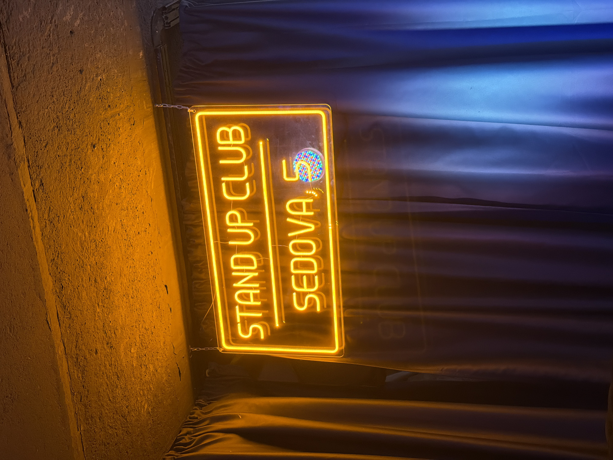 An illuminated neon sign hangs from a ceiling, displaying the text "STAND UP CLUB" on the top line and "SEDOVA, 5" on the bottom line. The "5" is accompanied by a circular graphic featuring multiple colored dots. The sign is framed by a yellow neon border. The background consists of dark curtains, with a visible gradient from purple on the left to blue on the right, illuminated by a blue light source. The ceiling above the sign appears to be a rough, concrete surface with an orange glow. The overall scene suggests the entrance or interior of a comedy club.