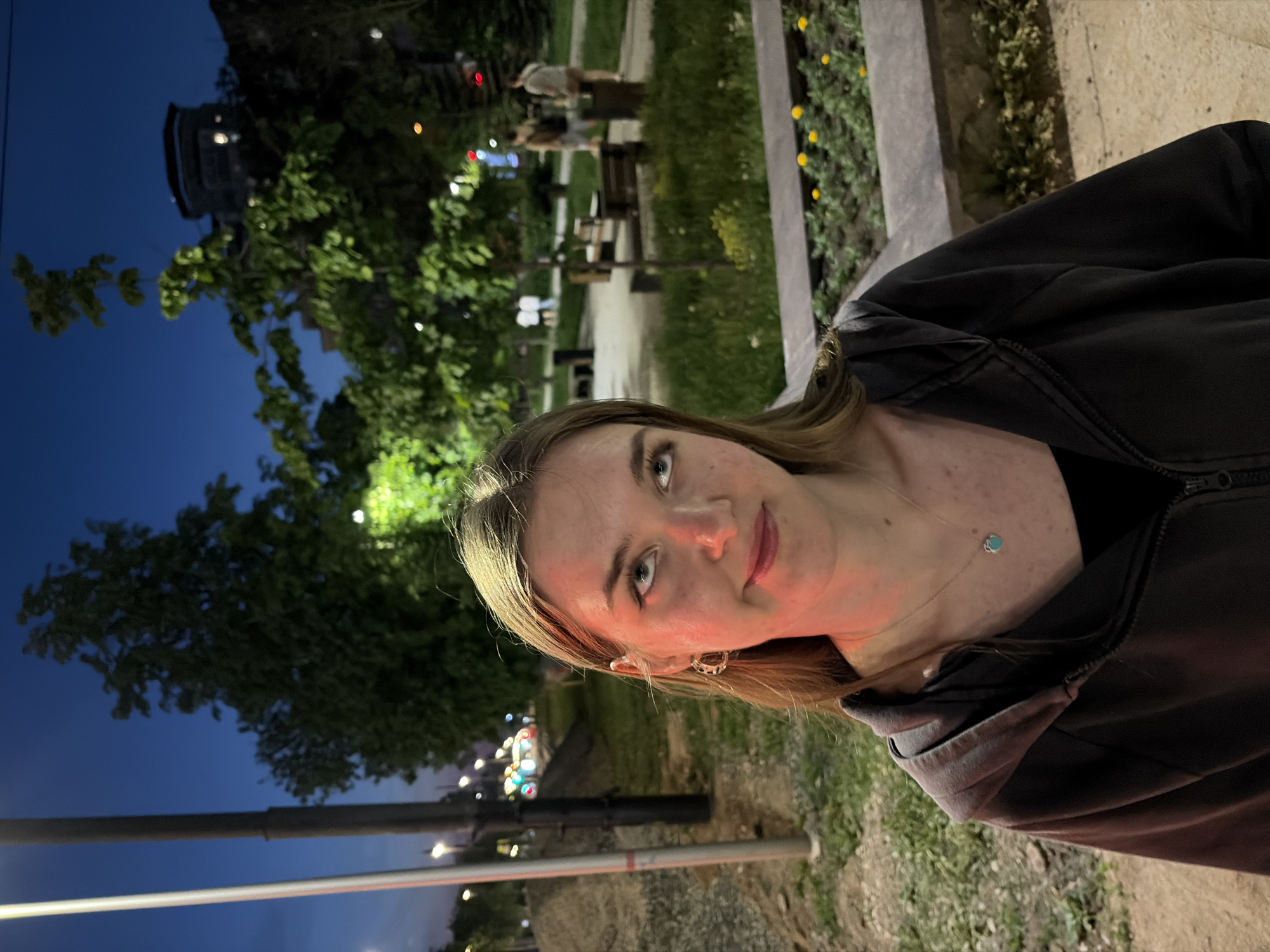 A woman with fair skin, light brown hair, and blue eyes is the primary subject of the photo, taken at night. She is wearing a dark hooded sweatshirt and a delicate necklace with a small turquoise pendant. In the background, there is a park-like area with trees, grass, and benches. Yellow flowers are planted along a border, and people can be seen indistinctly in the distance. Streetlights provide illumination. The location is Perm, Russia. A metal pole or railing runs horizontally in the foreground.