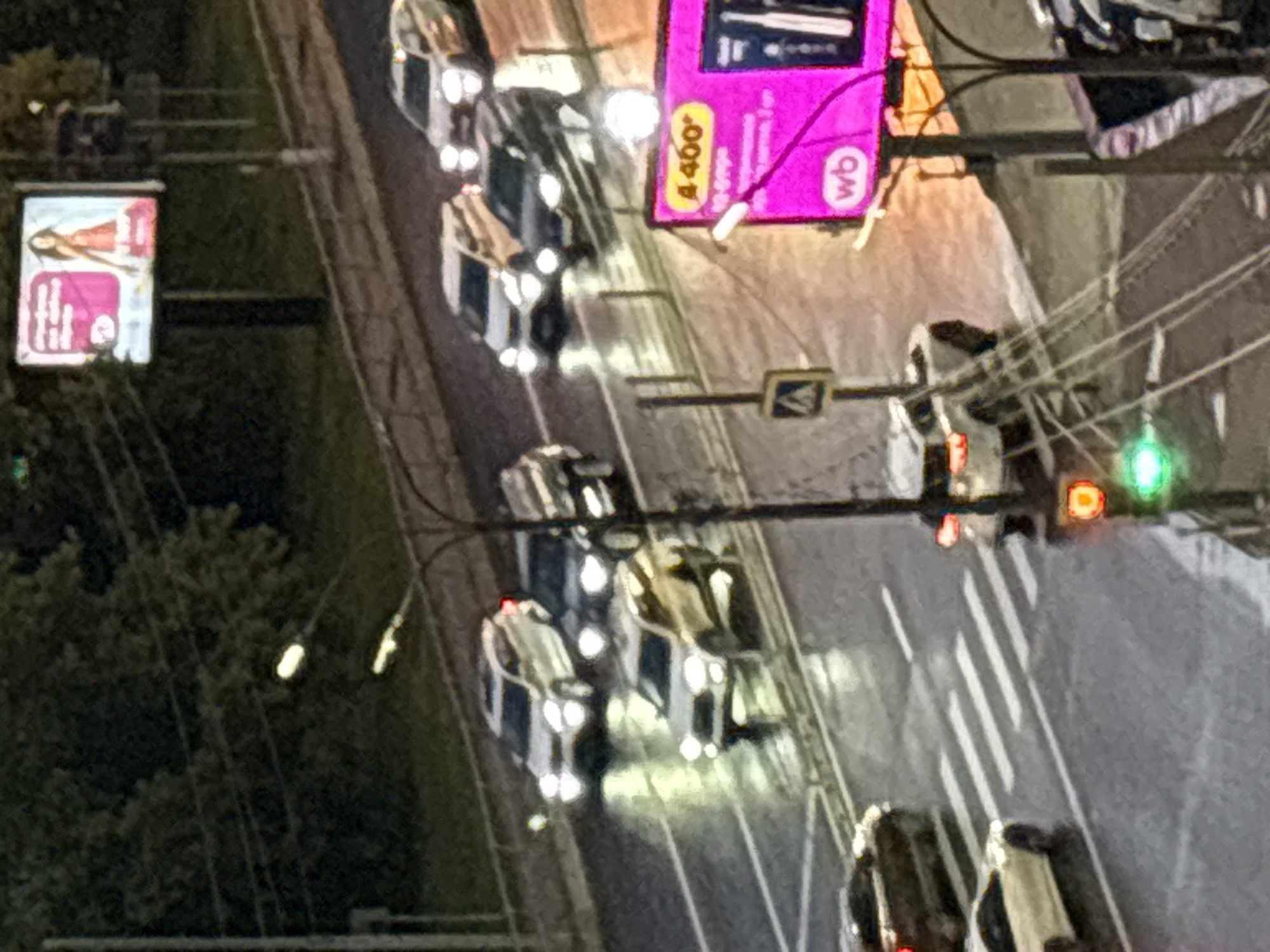 The image depicts a nighttime street scene in the city of Mytishchi, Russia, with several cars driving on a wet road, indicated by reflections. A billboard advertising is visible in the upper right, featuring a woman in a red dress. Traffic lights are present, with one showing a green light. A pedestrian crossing is also visible. The overall lighting suggests it is dusk or nighttime, with streetlights and car headlights illuminating the scene.
