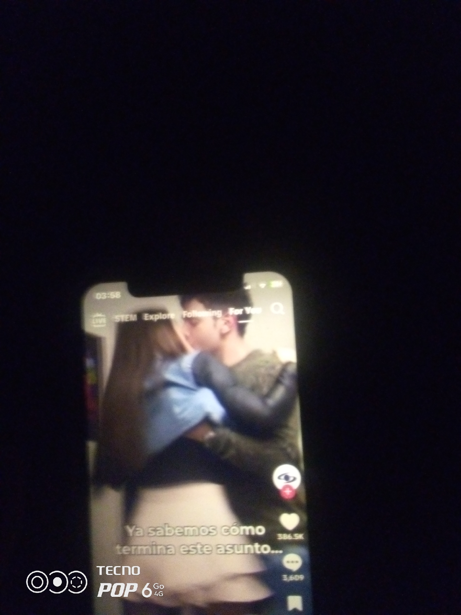 The image shows a smartphone screen displaying a TikTok video. The video features a couple kissing, with text overlayed that reads "Ya sabemos cómo termina este asunto..." which translates to "We already know how this matter ends...". The phone's interface is visible with options like "LIVE", "STEM", "Explore", "Following", and "For You". There are also icons for likes, comments, and shares. The bottom left corner of the phone displays "TECNO POP 6 4G". The image is dark, suggesting it was taken in low light conditions. There is no discernible indication of the location within Howayi, Nigeria.