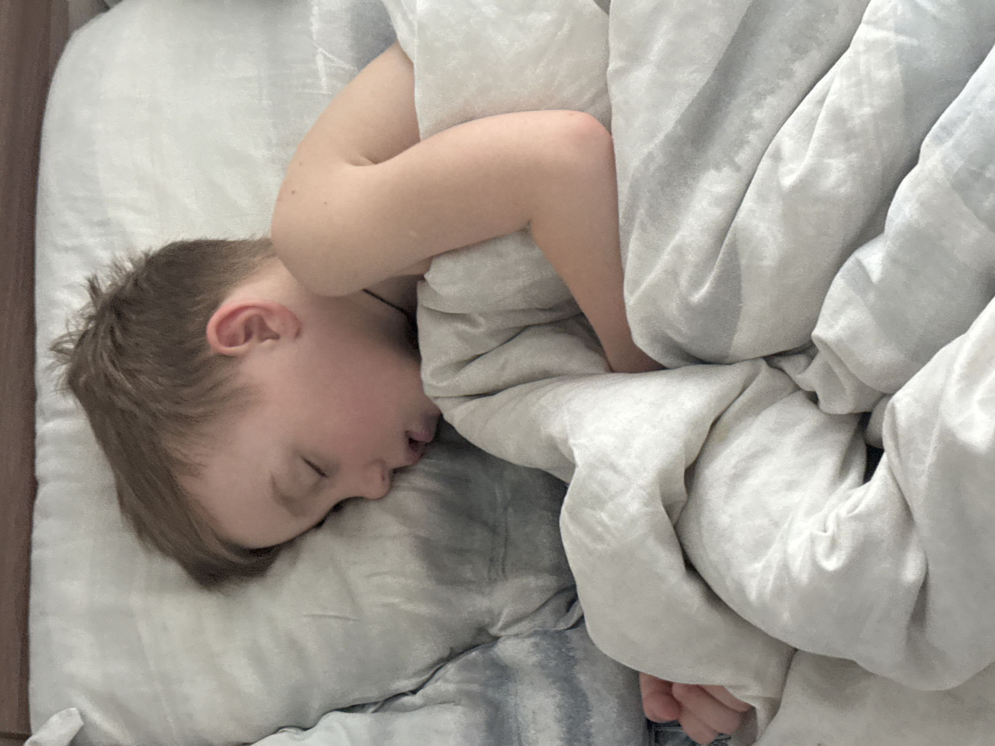 A young child is depicted sleeping peacefully in a bed. The child, who appears to be a male with light brown hair, is lying on their side with their head resting on a light grey or white pillow. Their eyes are closed and their mouth is slightly open, indicative of deep sleep. A soft, light grey or white duvet or comforter covers most of the child's body, appearing bunched and folded around them. One arm is partially visible, bent and tucked close to their chest, while the rest of their limbs are covered by the bedding. A darker grey or blue patterned sheet or blanket can be seen beneath the child's head and body. The setting is an indoor bedroom, suggesting a quiet moment, likely during the morning or nighttime. The overall atmosphere conveys a sense of tranquility and rest. No text is visible within the image. The image is oriented with the child's head to the left. The context for this image is Chaikovsky, Russia.