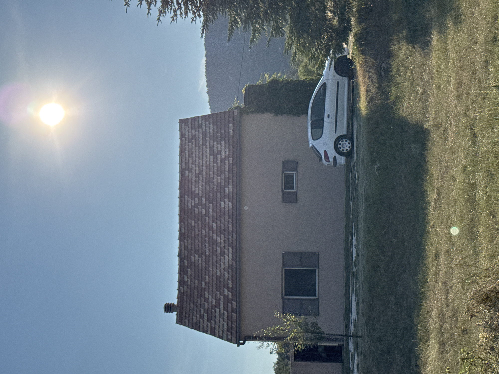 The image depicts a residential scene in Kopito, Montenegro, bathed in bright sunlight. In the foreground, a patch of dry grass and vegetation is visible. Behind this, a low mound of grass leads up to a paved area. A white compact car is parked on this paved surface to the right of a light-colored, two-story house with a red-tiled roof. The house has two windows with brown shutters, one larger and central, the other smaller and to the right. A chimney is visible on the roof. To the left of the house, a thin tree stands next to an open doorway. To the right of the house, a large evergreen tree is present. In the background, a hazy, dark mountain range is visible under a clear blue sky, with the sun prominently positioned in the upper right quadrant, casting a bright glare. The overall impression is of a rural or semi-rural residential property.