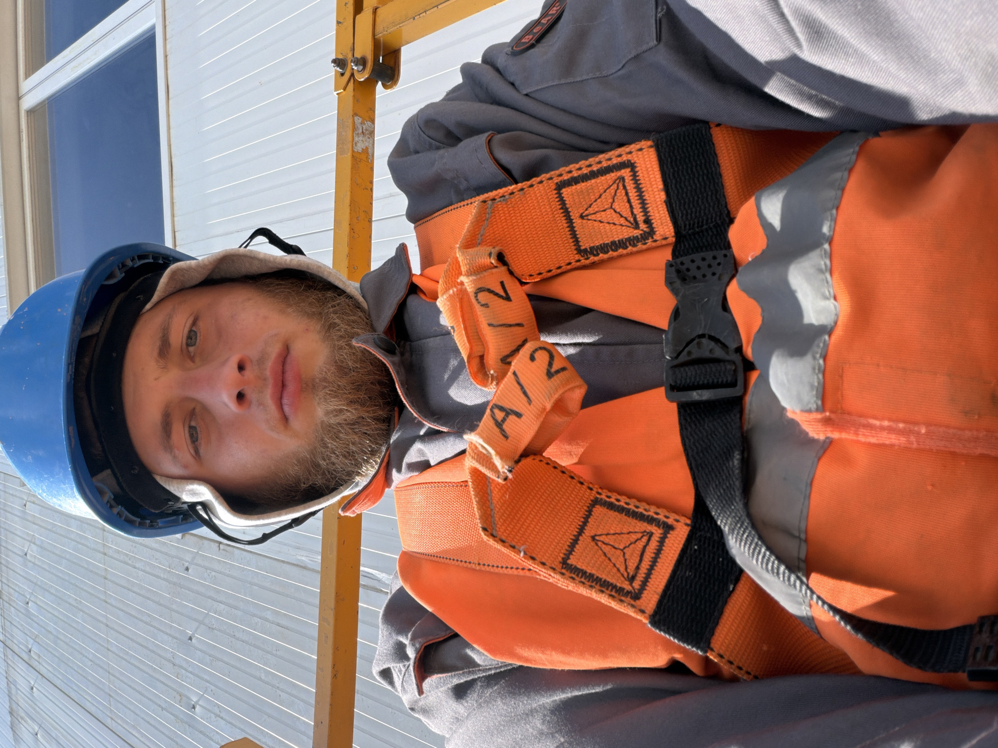 A close-up, low-angle, first-person perspective shot shows a man wearing a blue hard hat and an orange safety vest over a gray work uniform. He has a beard and is looking directly at the camera. The man is positioned in what appears to be an elevated work platform, with a yellow metal structure visible in the foreground. Behind him, a white corrugated metal wall and a large window are visible, suggesting an industrial or construction setting, likely in the vicinity of Stróża, Poland. The lighting indicates it is daytime, possibly outdoors.