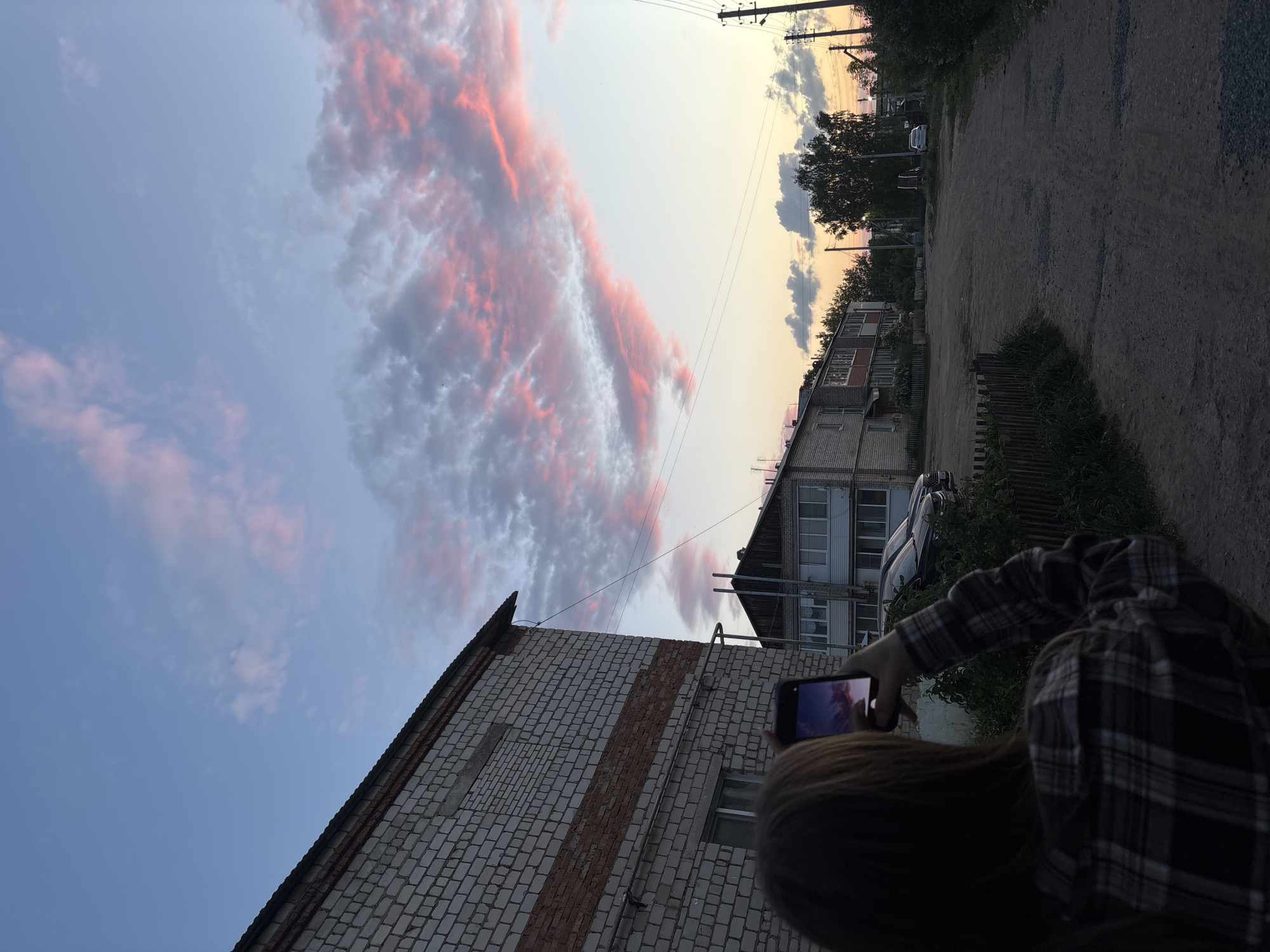 A vertical photograph depicts a person holding a smartphone, presumably taking a picture of a sunset in the city of Фоки, Russia. The sky is filled with pink-tinged clouds above low-rise buildings. A dirt road is to the right and power lines are strung above. The person taking the photo is facing away from the camera, and only the back of their head and arm are visible. The smartphone screen is reflecting the sunset scene.