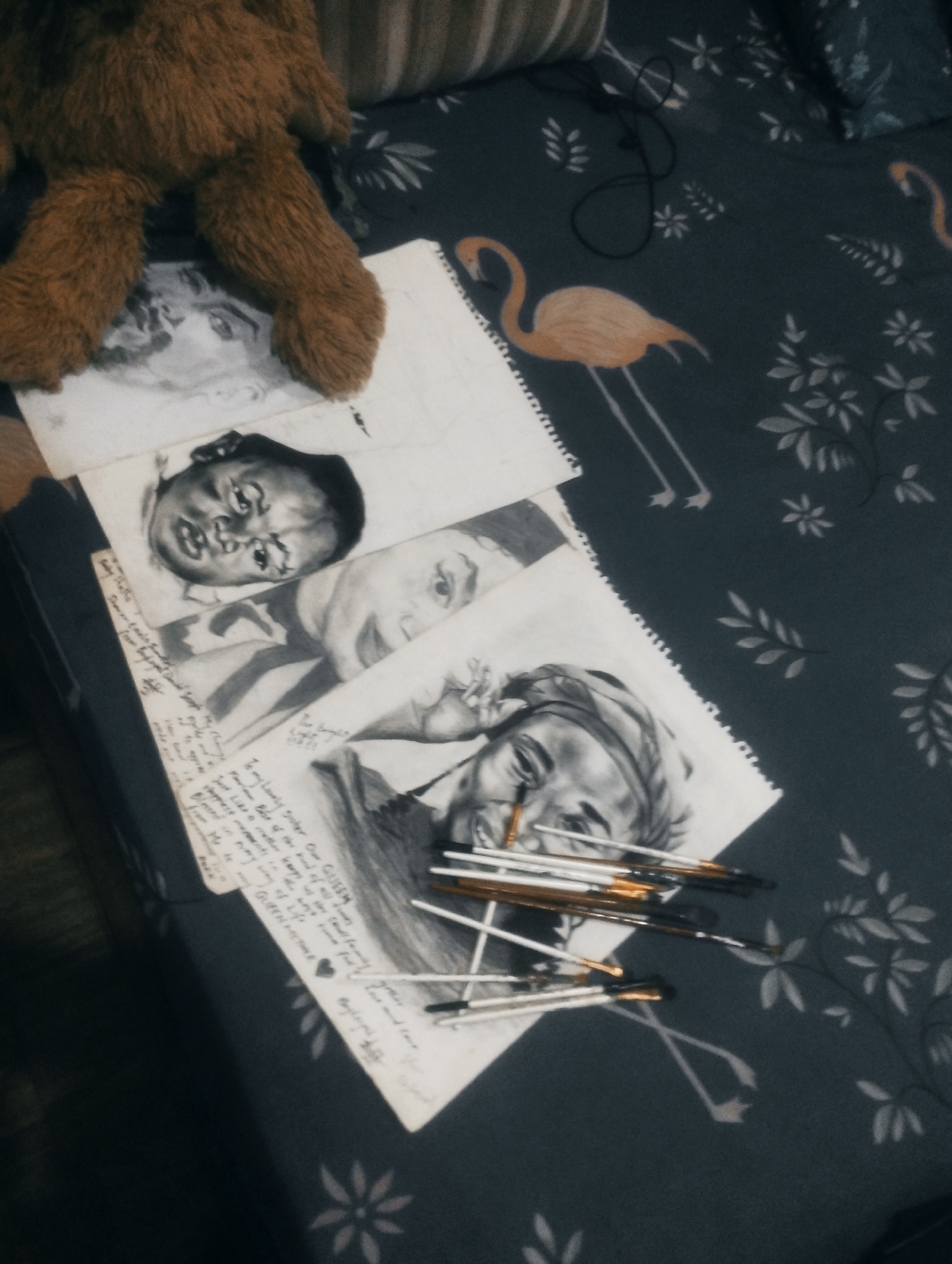 A close-up, high-angle shot depicts several charcoal sketches on paper laid out on a dark patterned fabric. The sketches are of human faces and appear to be unfinished. A teddy bear's furry legs are visible in the upper left corner, resting on one of the sketches. A collection of paintbrushes lies across the bottom of the frame, also resting on the sketches and fabric. The fabric has a repeating pattern of stylized white and orange flamingos and leaves against a dark background. A black string is loosely coiled near the top right of the image. The scene is dimly lit. The location is Unknown, Unknown.
