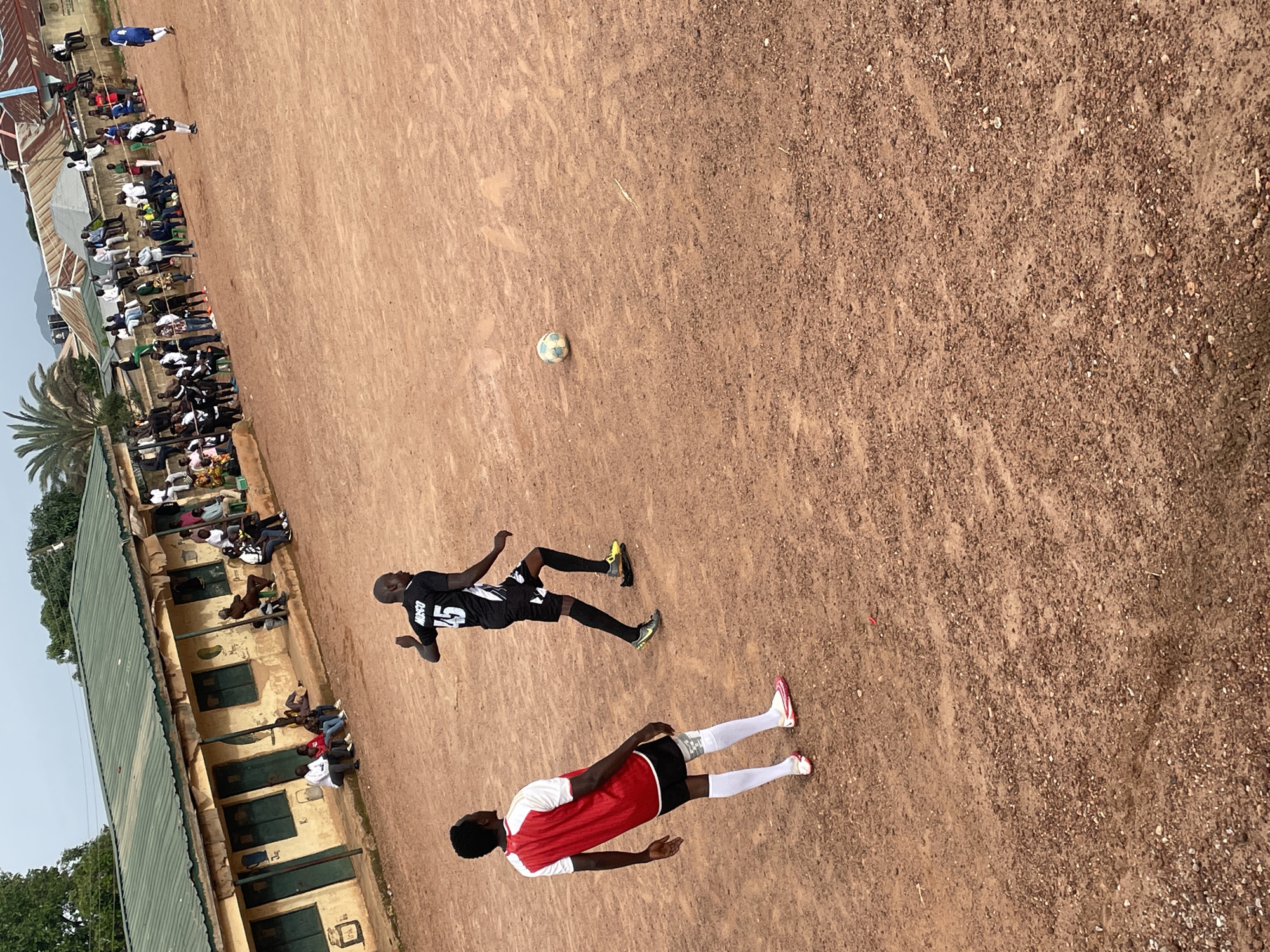 The image depicts a dusty outdoor football pitch in Jos, Nigeria, with a game in progress. Two male players are the main subjects, one in a red and white jersey and black shorts, the other in a black jersey with the number 45 and black shorts. A football lies on the ground between them. Spectators are seated and standing along a raised embankment in the background, with a low-rise building with a green roof visible behind them. The ground is a reddish-brown dirt, characteristic of many football pitches in Nigeria. The sky is overcast.