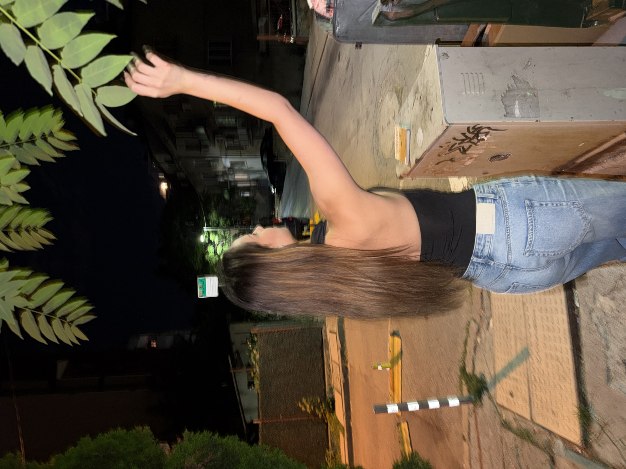 In this nighttime image, a woman with long, brown hair is shown from the back and side, wearing a black top and light blue jeans. She stands on a paved sidewalk next to a street, reaching up with her right hand to touch the leaves of a tree. To her right, a metal utility box, possibly an electrical cabinet, is present. In the background, buildings and a street are visible, illuminated by streetlights. The scene appears to be located in a residential area of Sofia, Bulgaria, as suggested by the street sign which reads "GREEN ZONE" in English and "ЗЕЛЕНА ЗОНА" in Bulgarian.