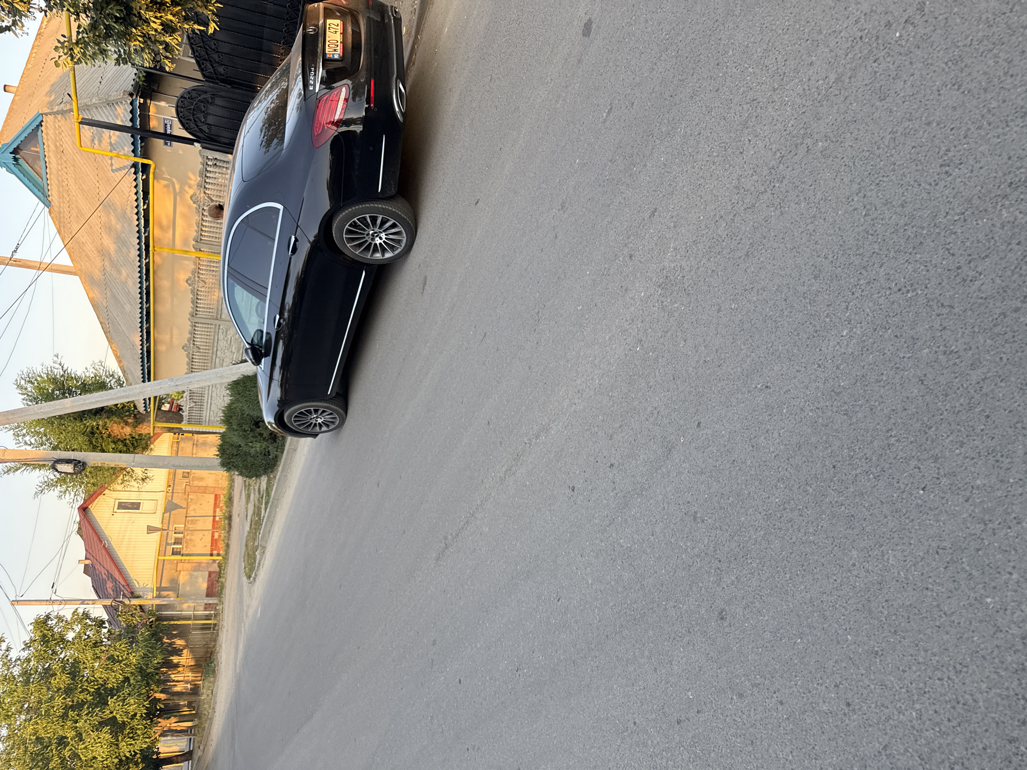 The image shows a black Mercedes-Benz E-Class parked on a street in Cahul, Moldova. The car is facing left, and the rear of the vehicle, including the license plate and taillights, is visible. The license plate reads "MDQ 472." A gas pipe runs along the side of a building behind the car, and a portion of the building's roof is visible. The street is paved with asphalt. In the background, there are houses and trees. The scene appears to be set during the day, possibly late afternoon or early evening, given the warm lighting and shadows. The weather appears to be clear.